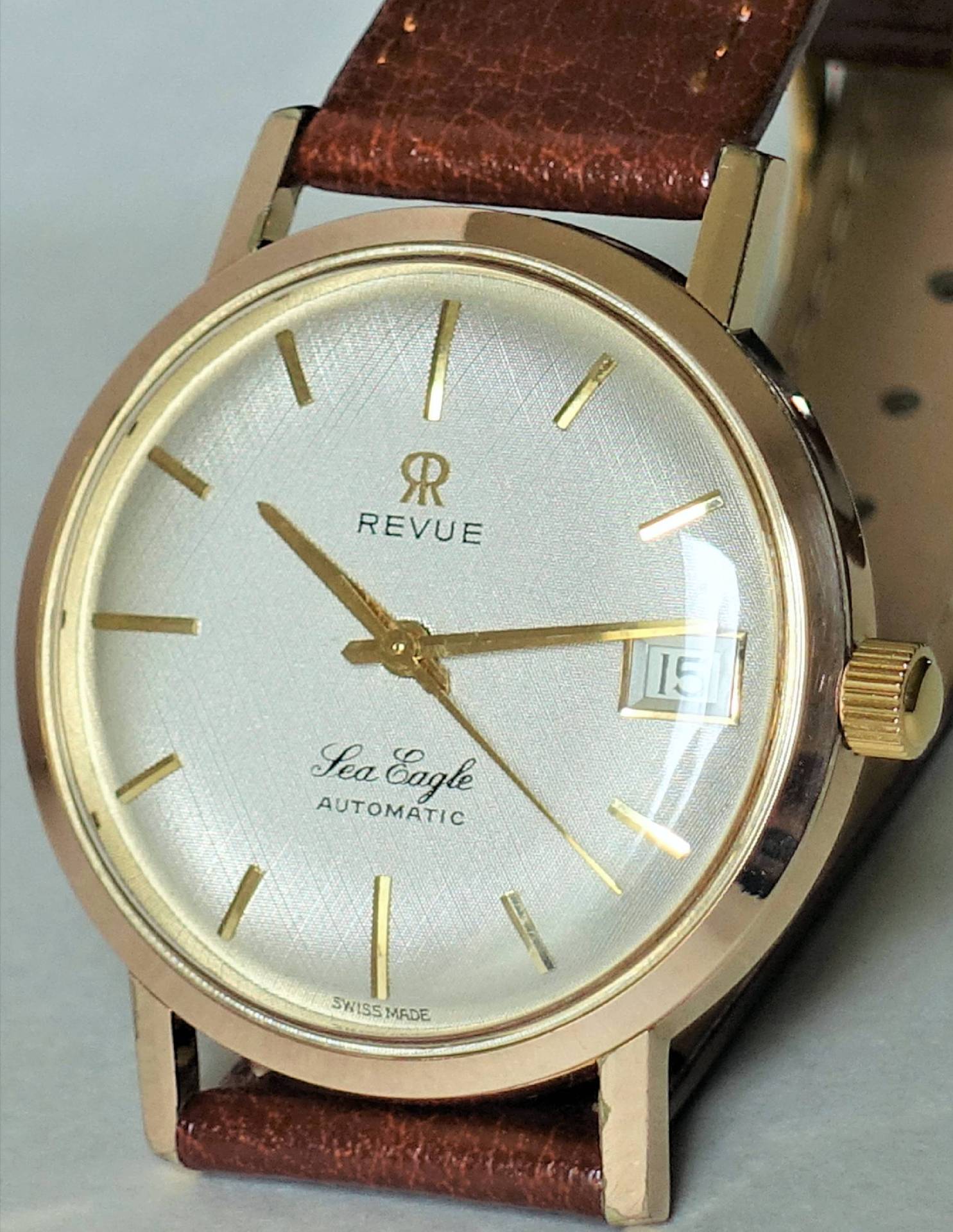 Goldfarbene Armbanduhr mit weißem Zifferblatt, braunem Armband und Datumsanzeige.
