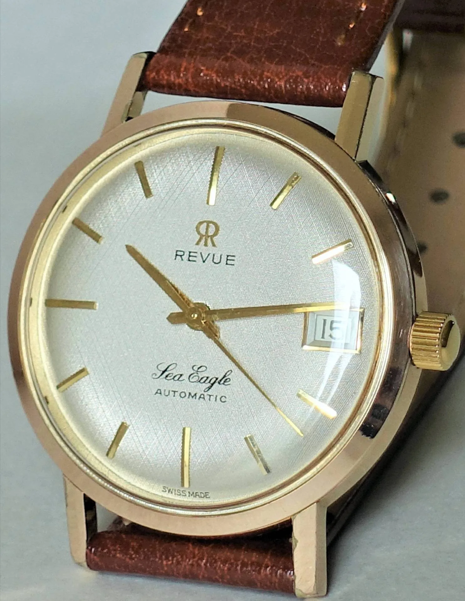 Goldfarbene Armbanduhr mit weißem Zifferblatt, braunem Armband und Datumsanzeige.