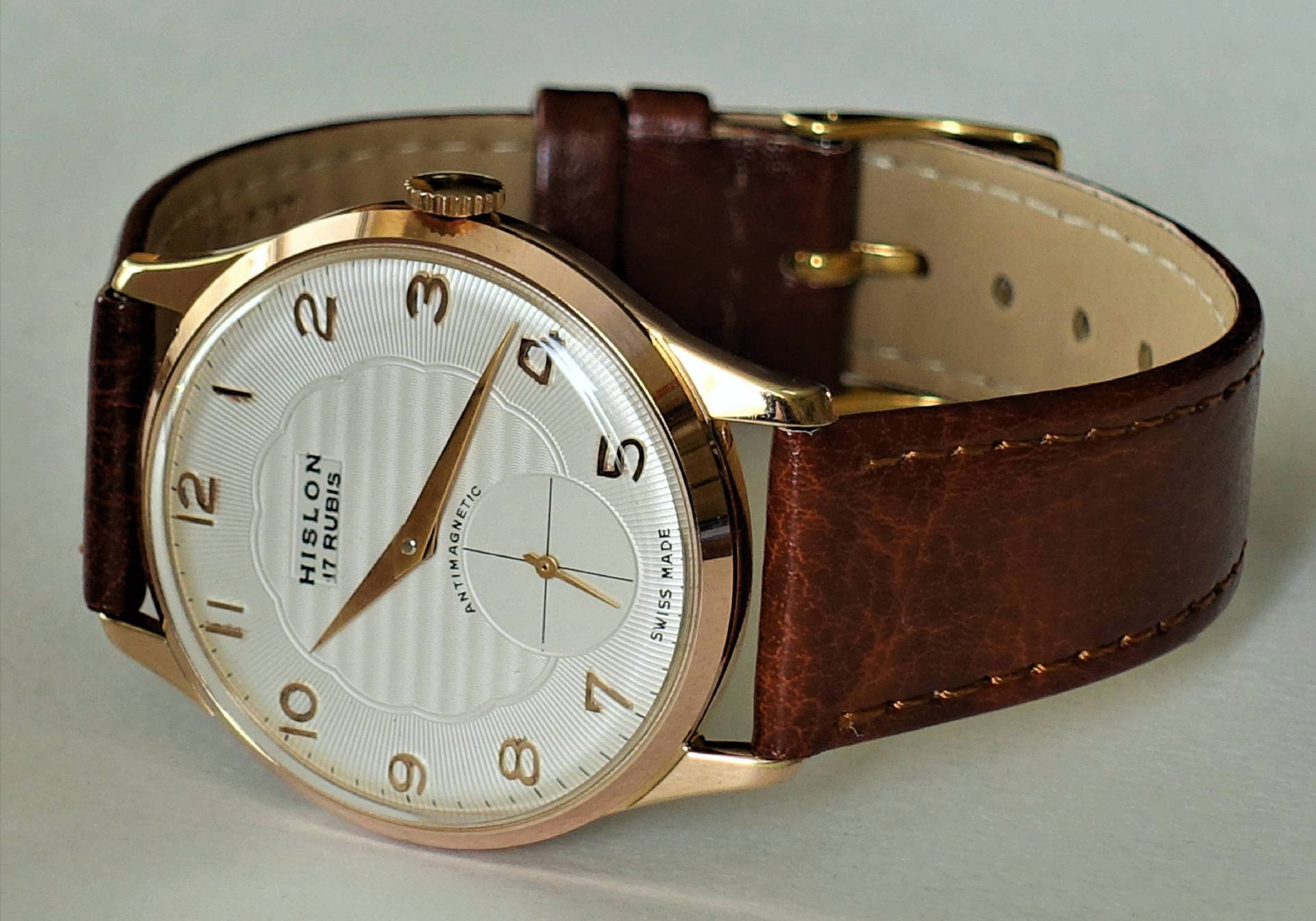 Eine Armbanduhr mit weißem Zifferblatt, goldfarbenem Gehäuse und braunem Lederarmband liegt auf einer hellen Oberfläche.