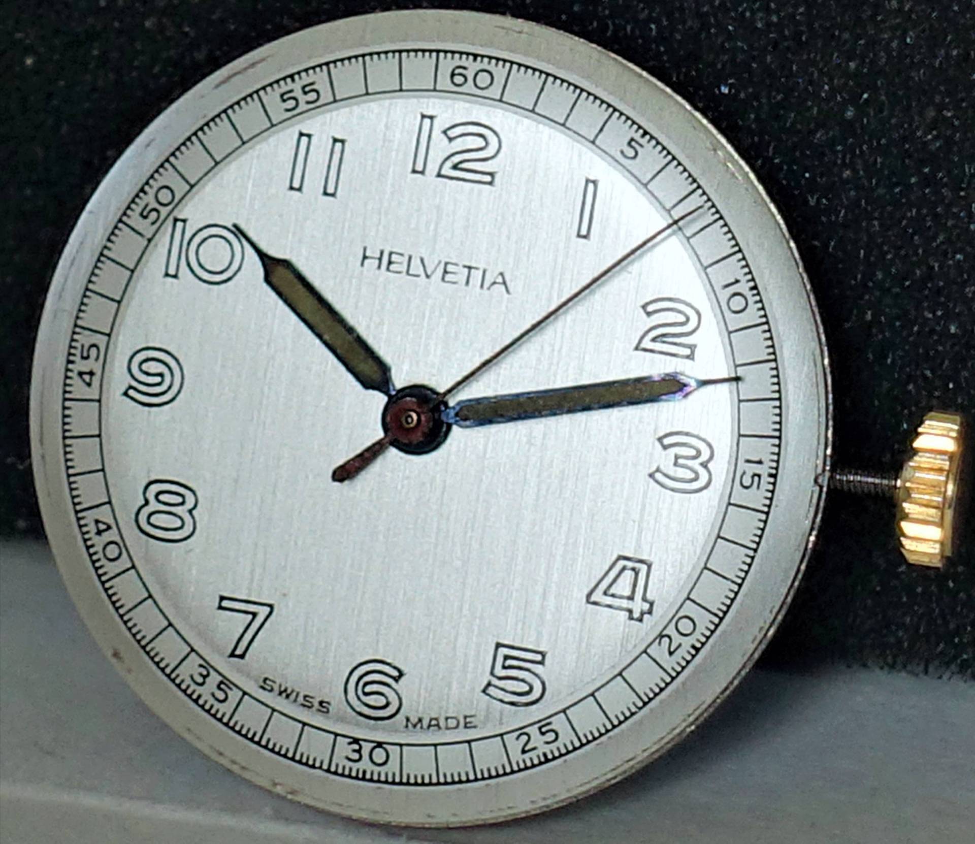 Silberfarbenes Helvetia Uhrwerk mit Zifferblatt, Zeigern und goldfarbener Krone.