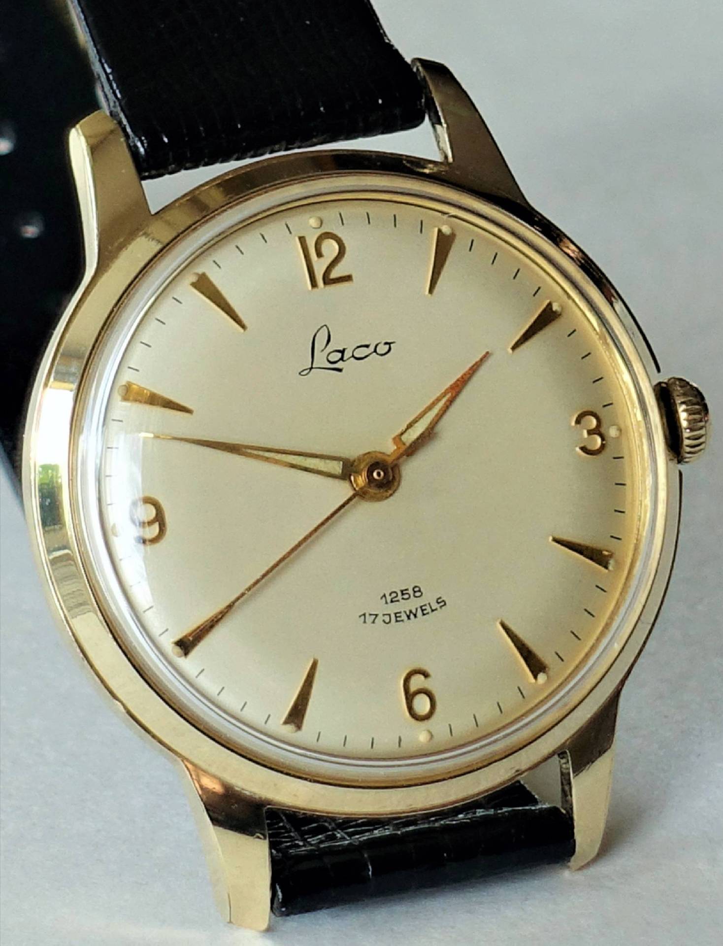 Goldfarbene Armbanduhr mit hellem Zifferblatt, schwarzen Zahlen und schwarzem Armband.