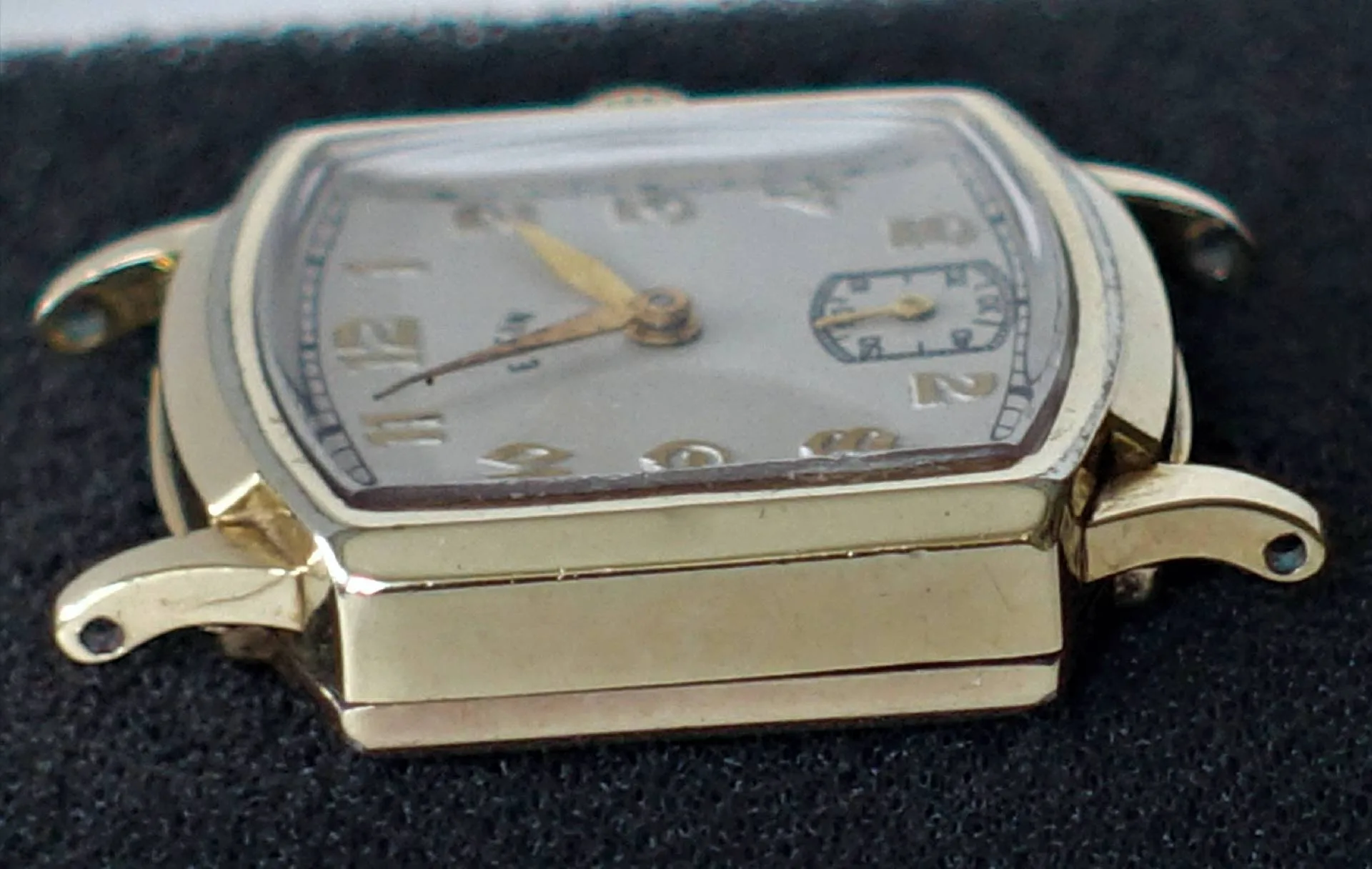 Eine rechteckige, goldfarbene Armbanduhr mit silberfarbenem Zifferblatt und arabischen Zahlen liegt auf schwarzem Stoff.