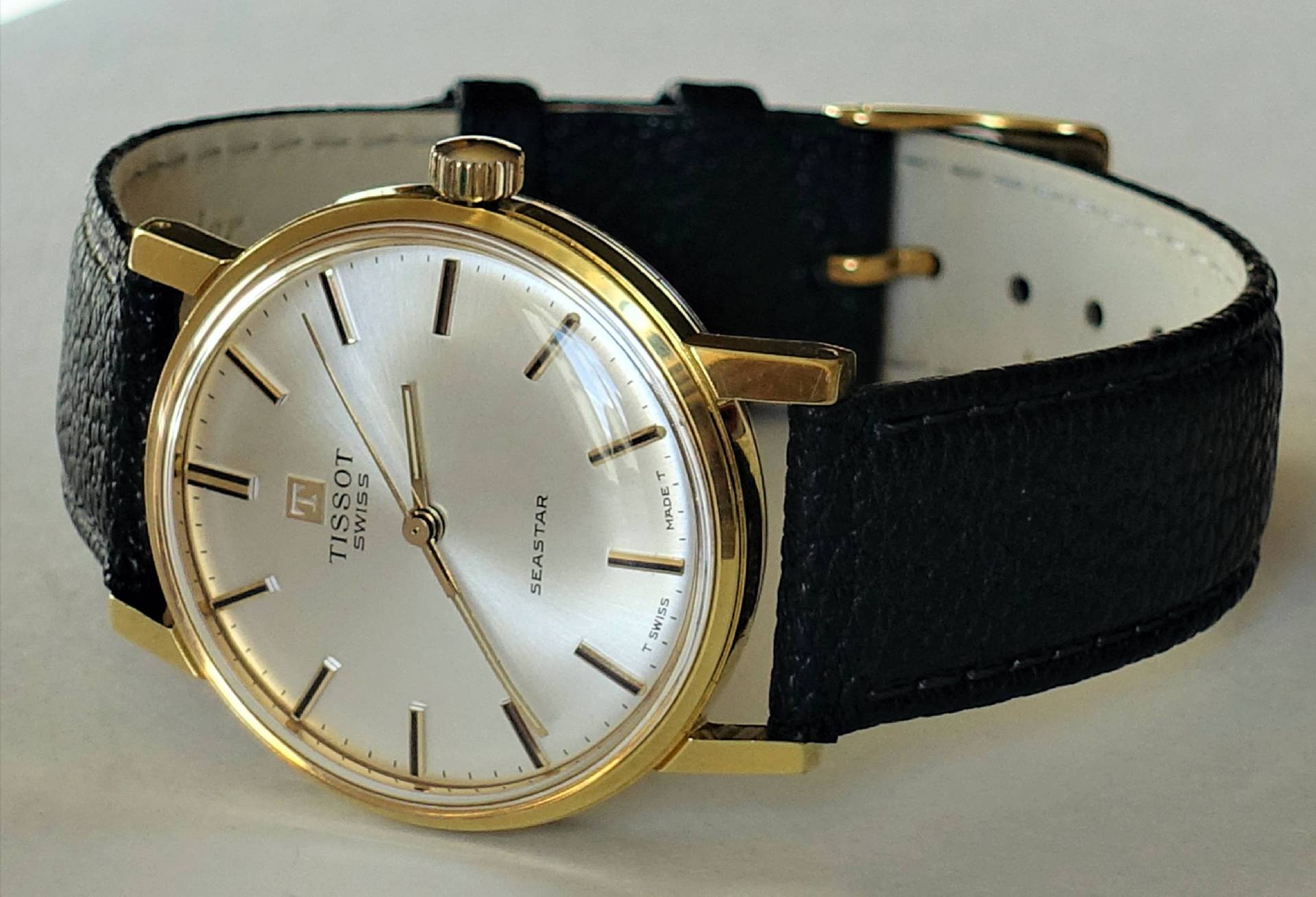 Klassische Armbanduhr mit goldfarbenem Gehäuse, silberfarbenem Zifferblatt und schwarzem Armband.