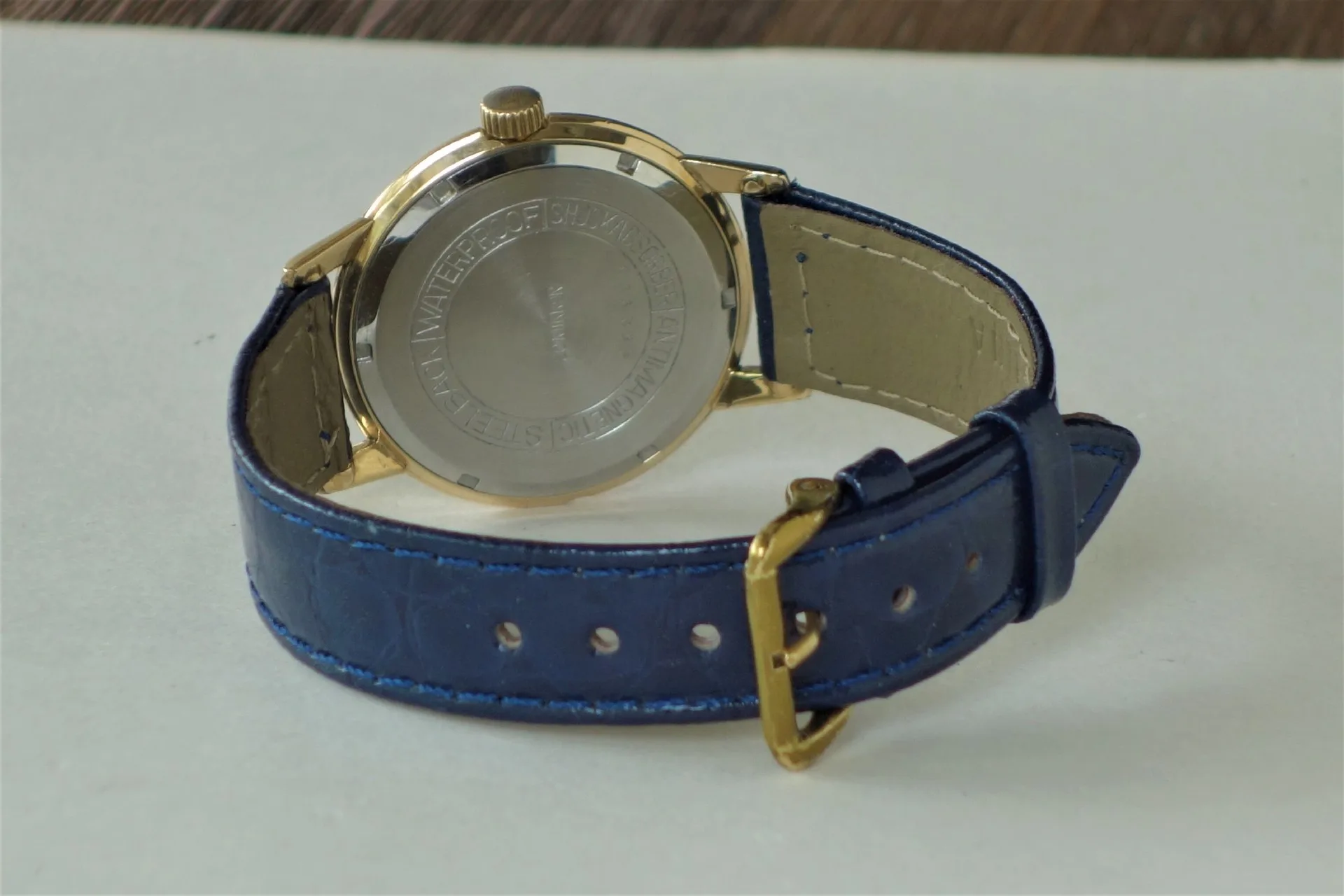 Armbanduhr mit blauem Armband und goldfarbenem Gehäuse, Rückseite sichtbar.