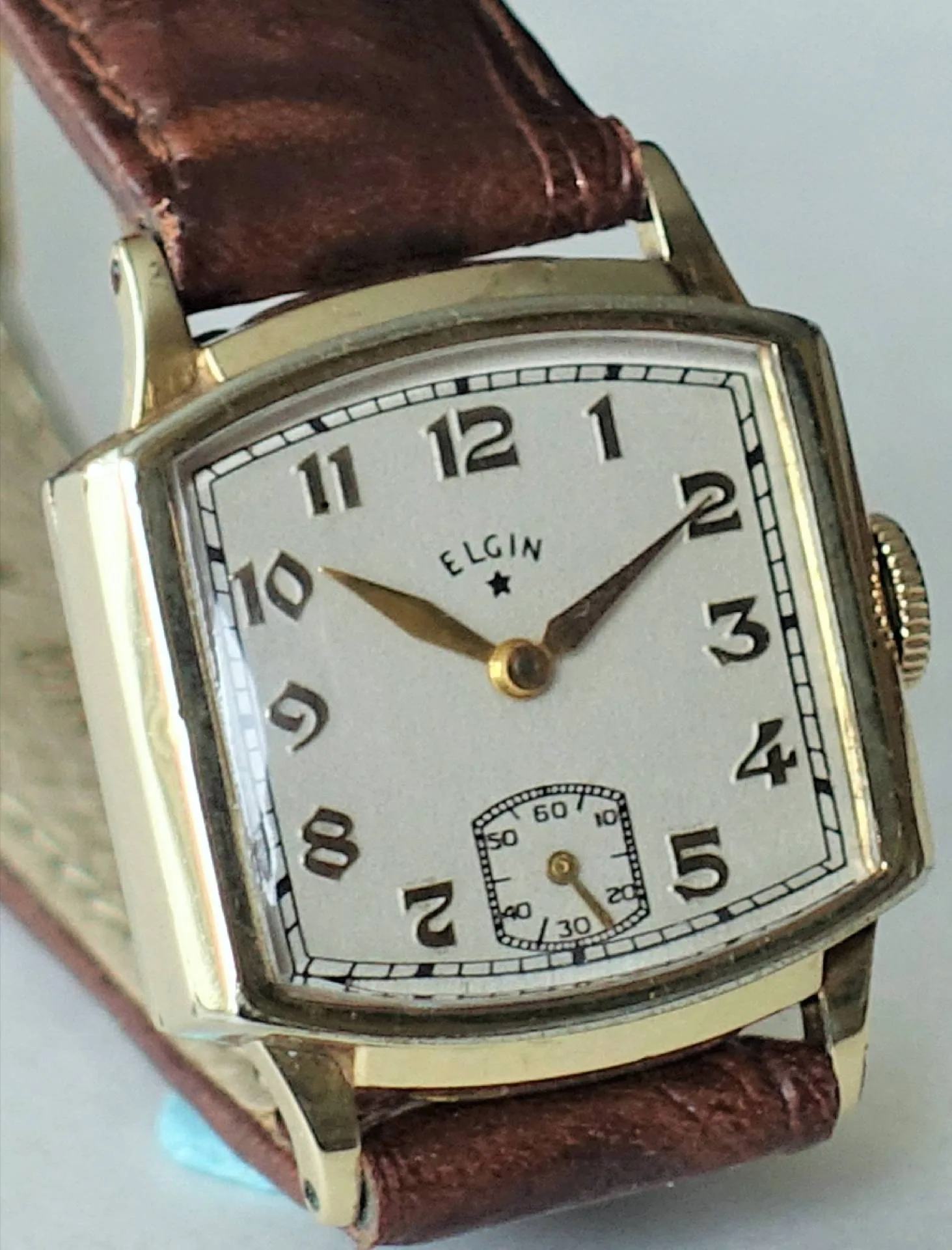 Eine rechteckige Armbanduhr mit gelbfarbenem Gehäuse, weißem Zifferblatt, arabischen Zahlen und braunem Lederarmband.