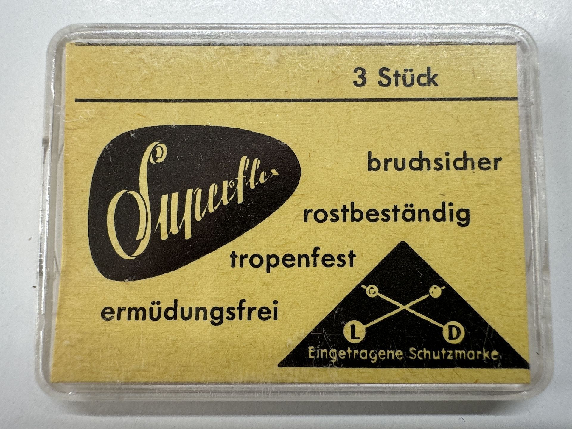 Gelbe Verpackung mit drei Federstegen für Uhren, Aufschrift "Superflex" und Symbol mit zwei gekreuzten Uhrzeigern.