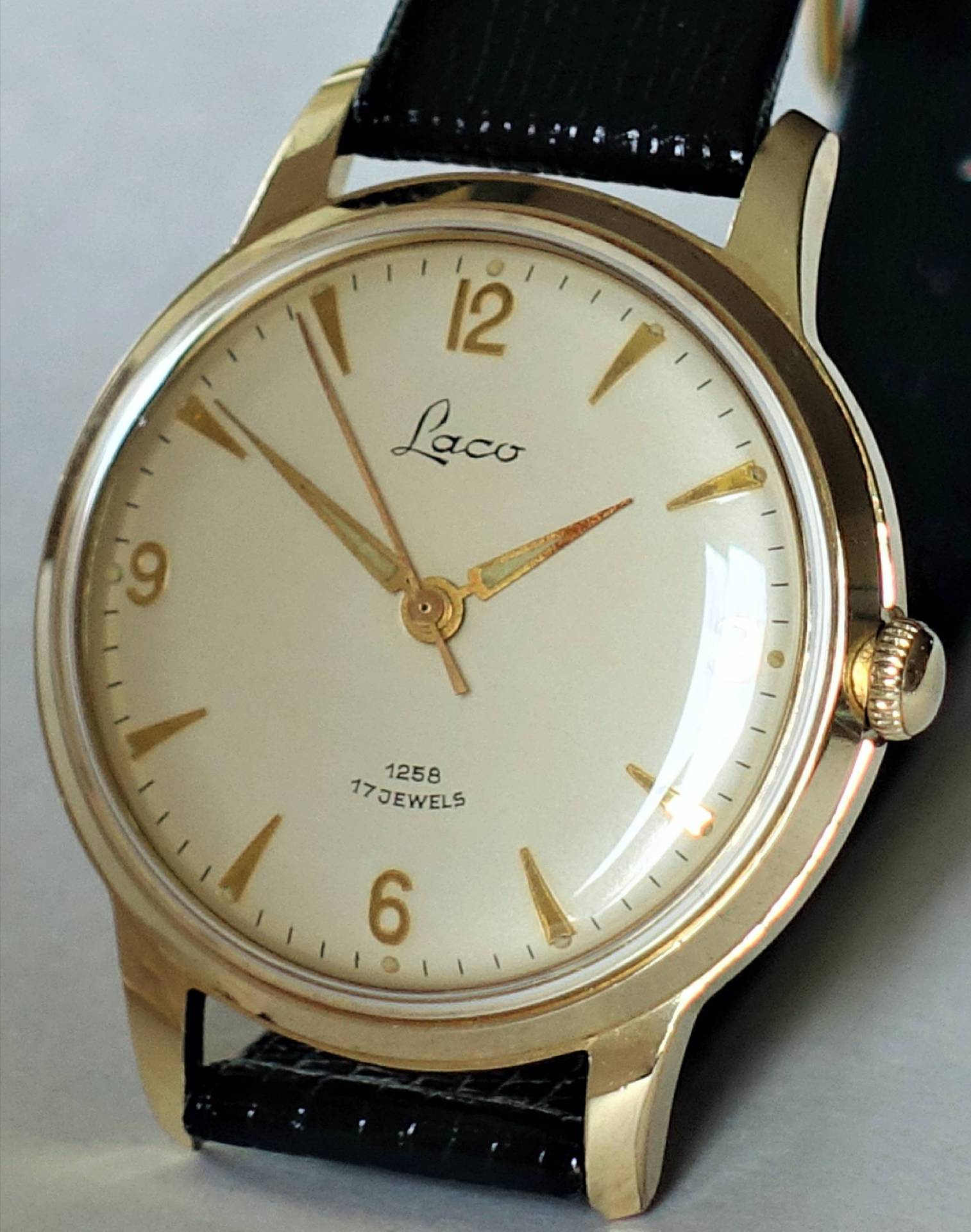 Goldfarbene Armbanduhr mit hellem Zifferblatt, goldenen Indizes und schwarzem Armband.