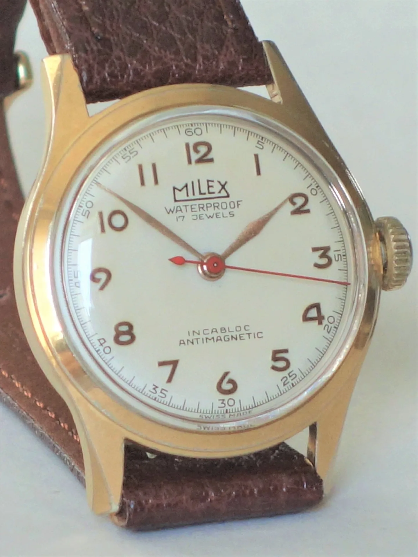Analoge Armbanduhr der Marke Milex mit goldfarbenem Gehäuse, weißem Zifferblatt und braunem Armband.