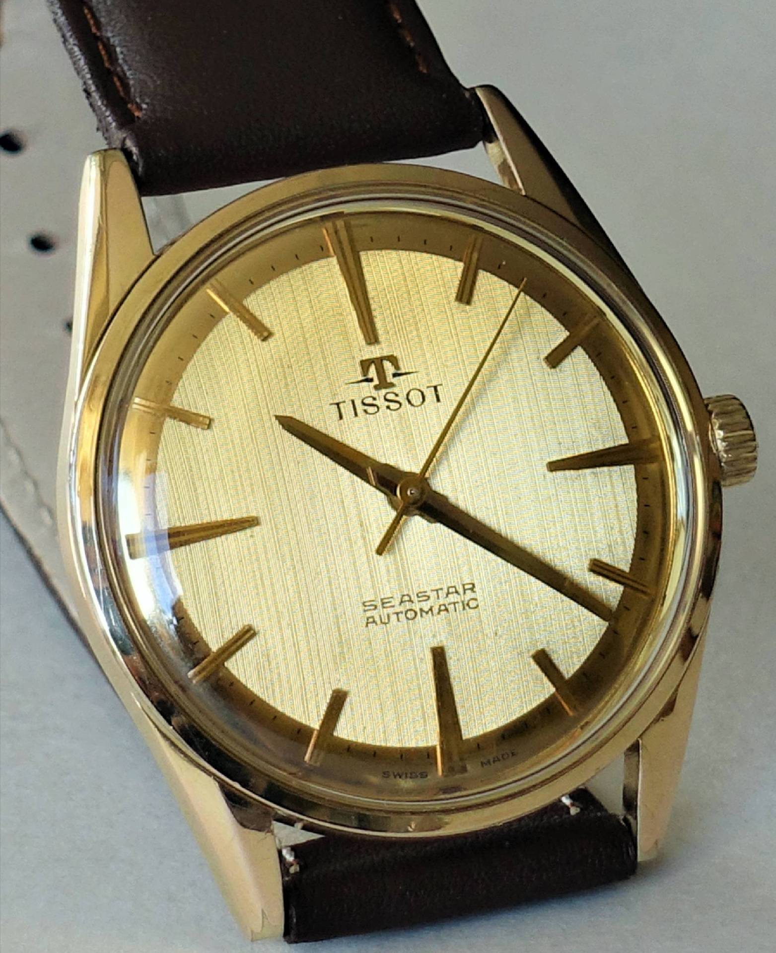Goldfarbene Armbanduhr mit hellem Zifferblatt und braunem Armband.