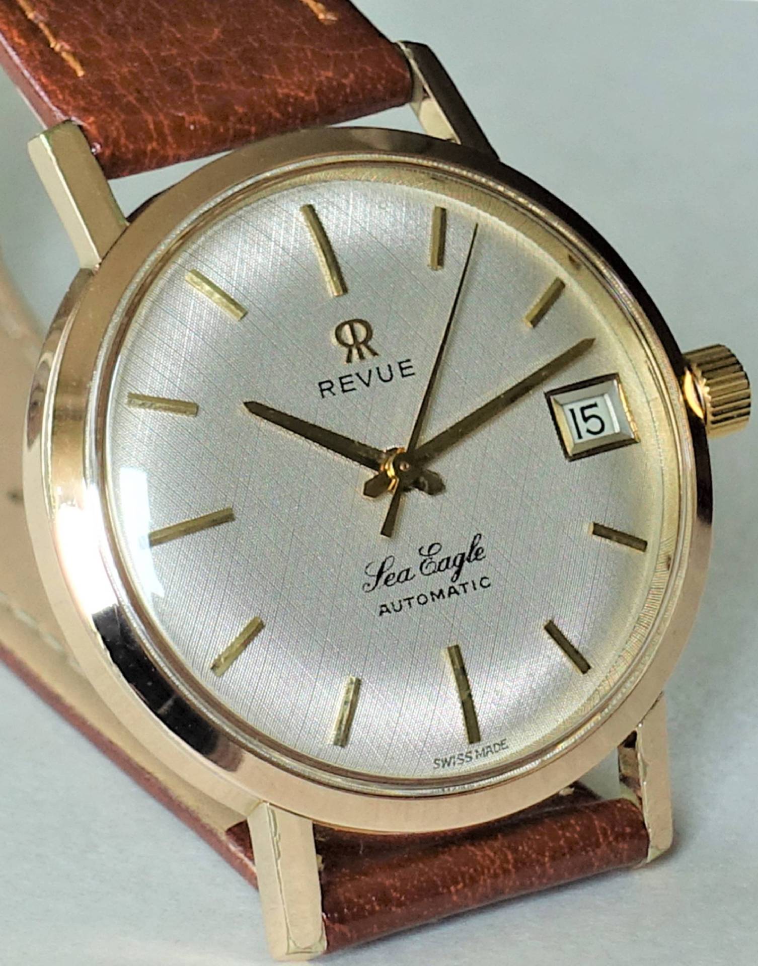 Goldfarbene Armbanduhr mit silbernem Zifferblatt, braunem Armband und Datumsanzeige.
