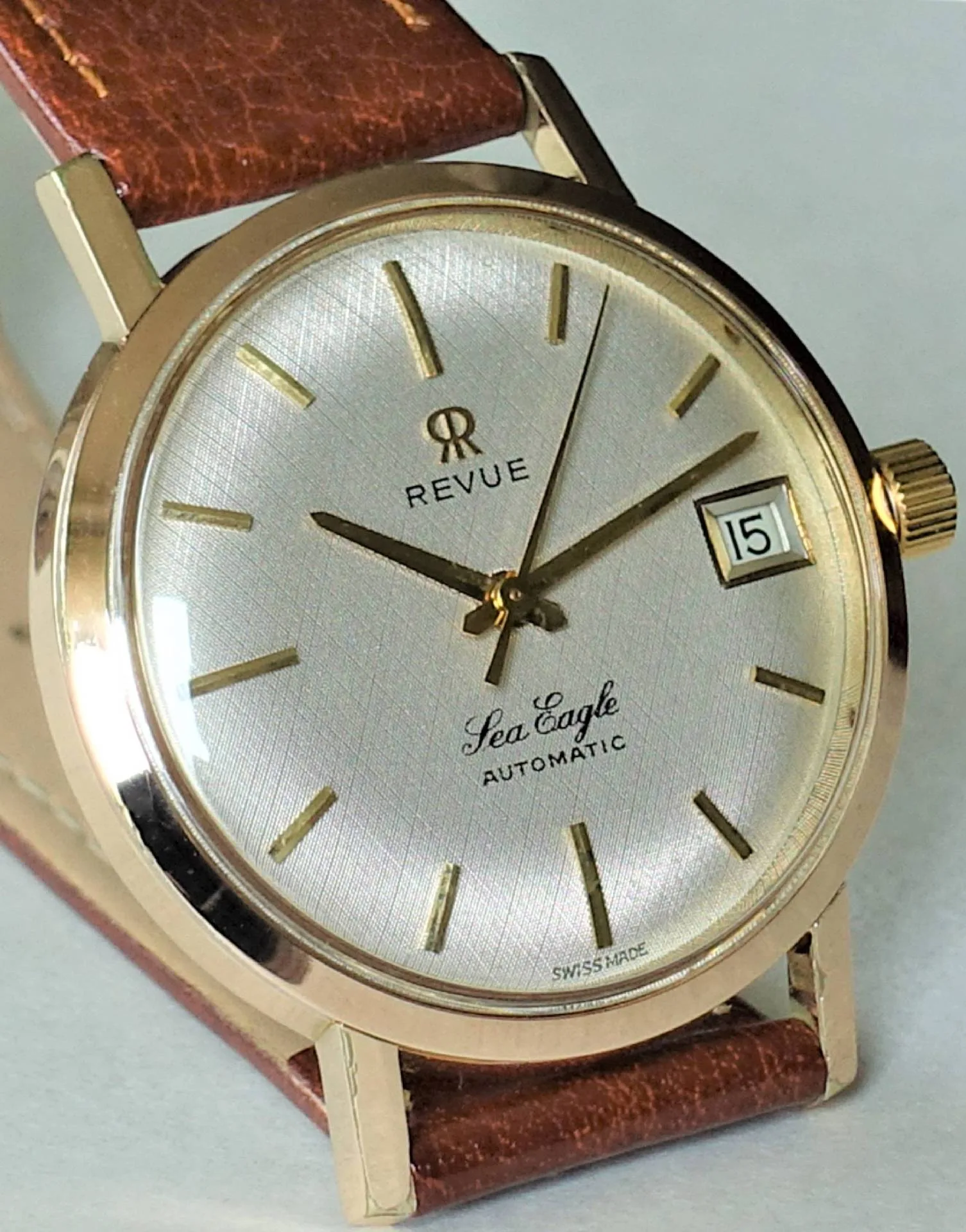Goldfarbene Armbanduhr mit silbernem Zifferblatt, braunem Armband und Datumsanzeige.