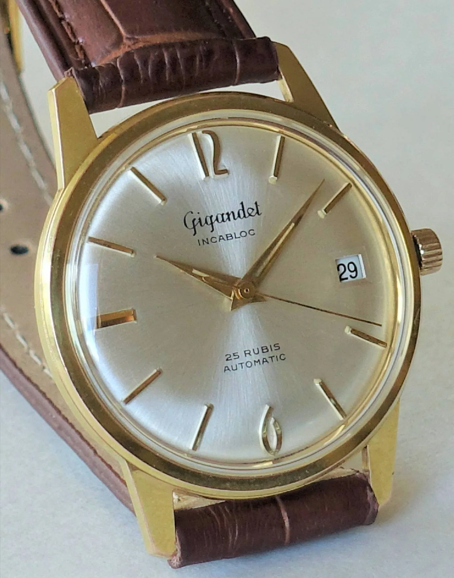 Eine klassische Armbanduhr mit goldfarbenem Gehäuse, silbernem Zifferblatt, braunem Lederarmband und Datumsanzeige bei drei Uhr.