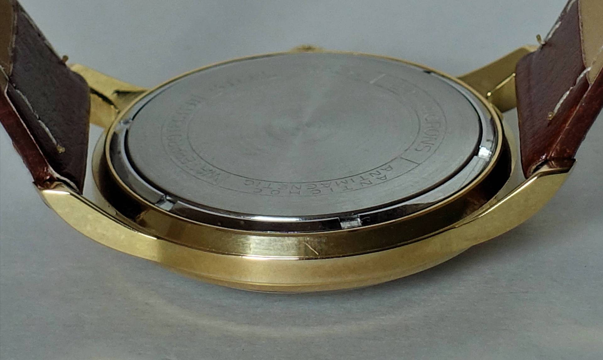 Silberfarbener runder Boden einer Armbanduhr mit goldfarbenem Gehäuse und braunem Armband.