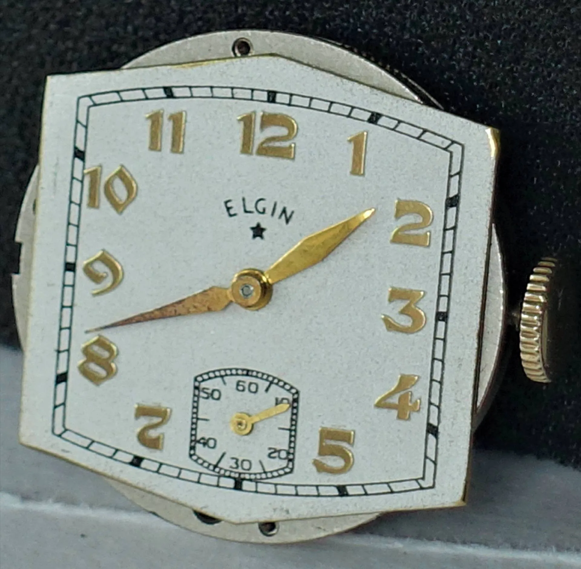 Weiß-goldenes eckiges Uhrwerk Das Bild zeigt ein eckiges Uhrwerk mit weißem Zifferblatt, goldfarbenen Zahlen und Zeigern sowie einer kleinen Sekunde bei sechs Uhr.