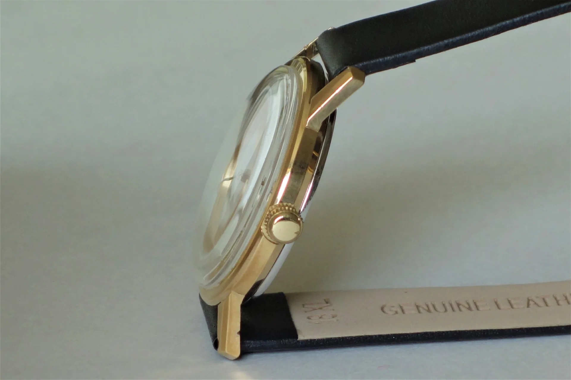Flache Armbanduhr mit goldfarbenem Gehäuse und schwarzem Lederarmband in Seitenansicht.