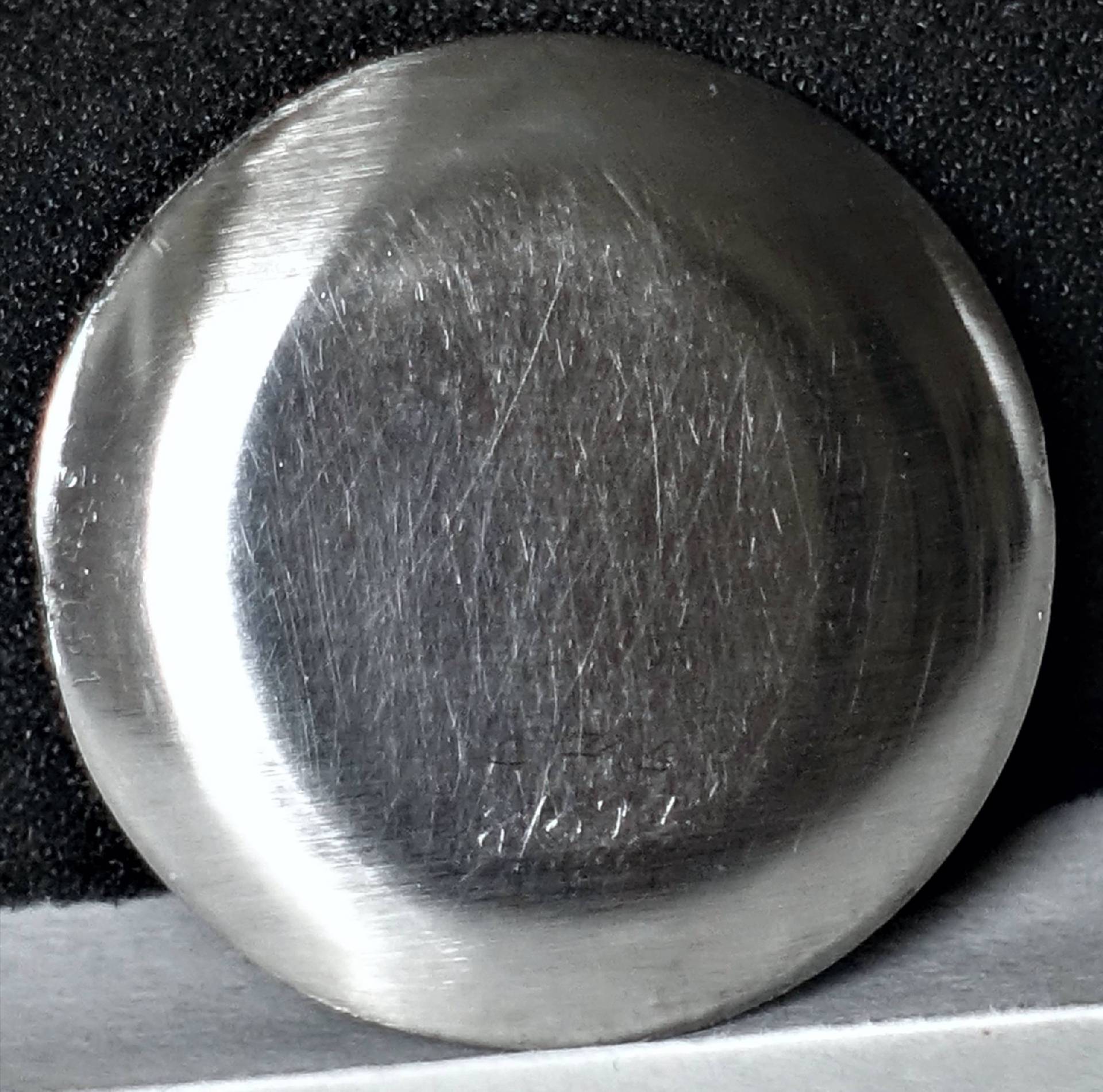 Eine silberfarbene, runde Rückseite eines Uhrendeckels mit sichtbaren feinen Kratzern liegt auf schwarzem Untergrund.