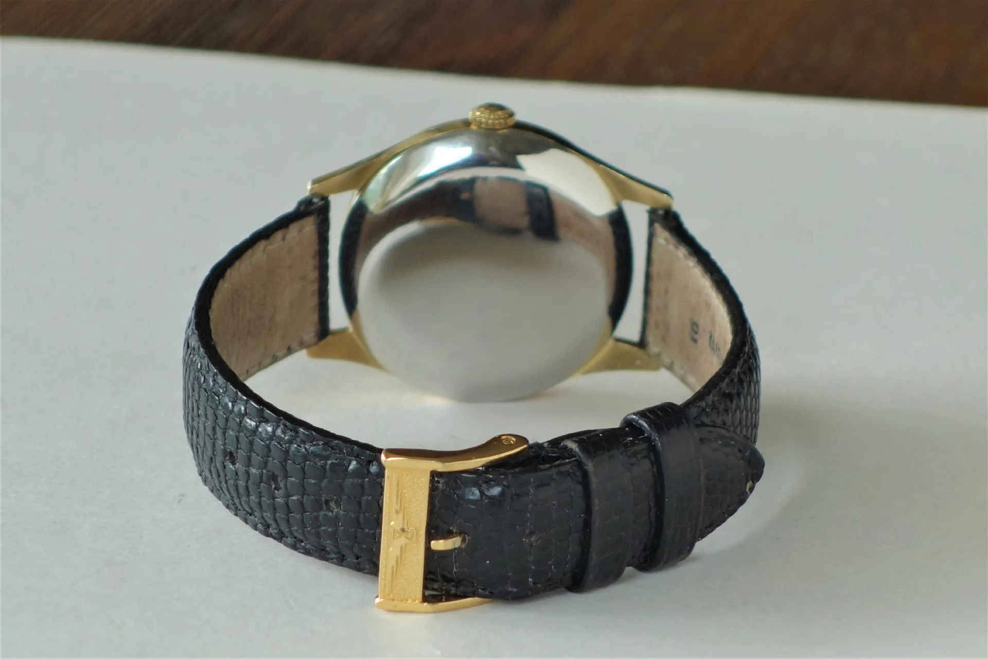 Armbanduhr mit schwarzem Armband und goldfarbenem Gehäuse, Rückseite sichtbar.