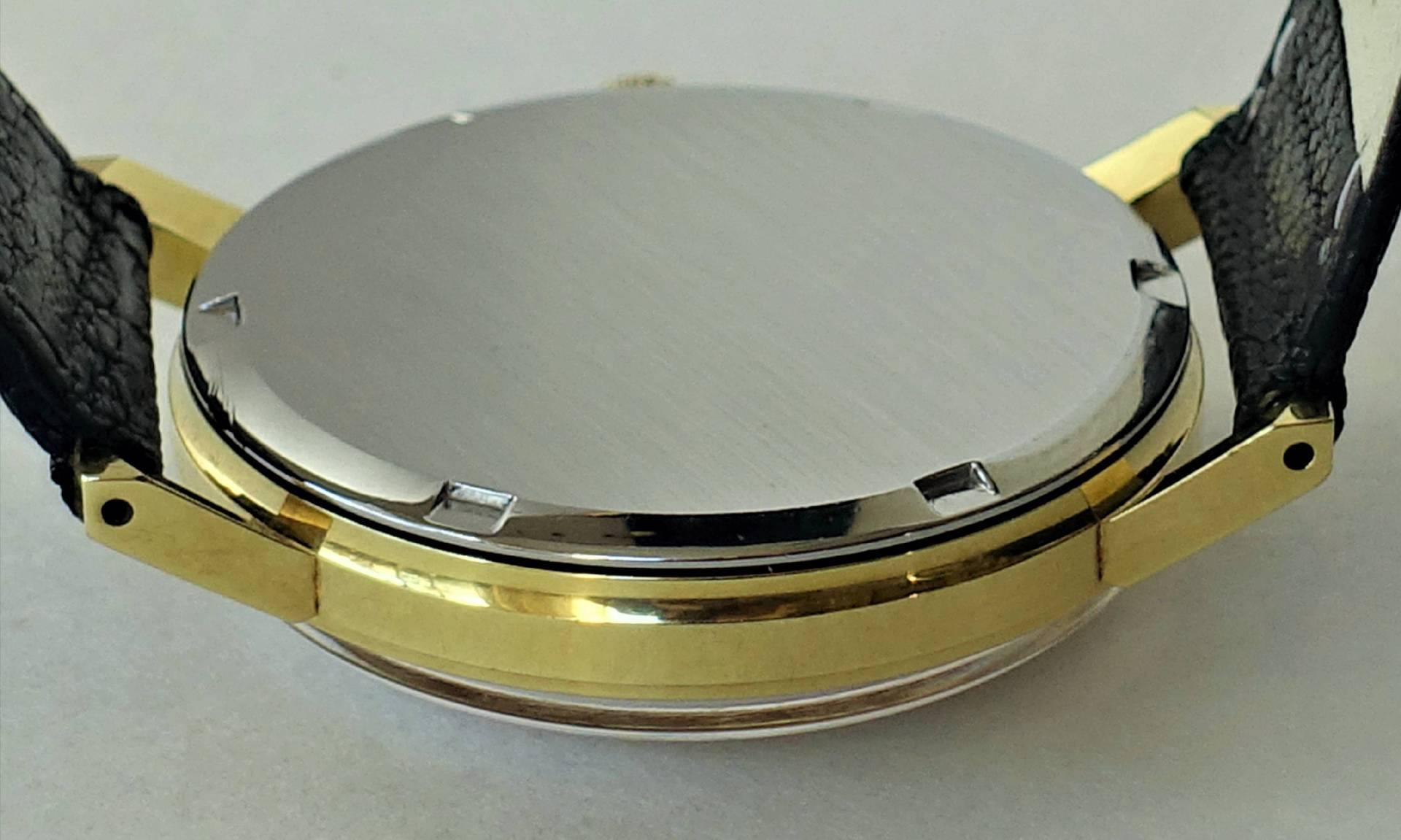 Silberfarbener Rückdeckel einer Armbanduhr mit goldfarbenem Gehäuse und schwarzem Armband.