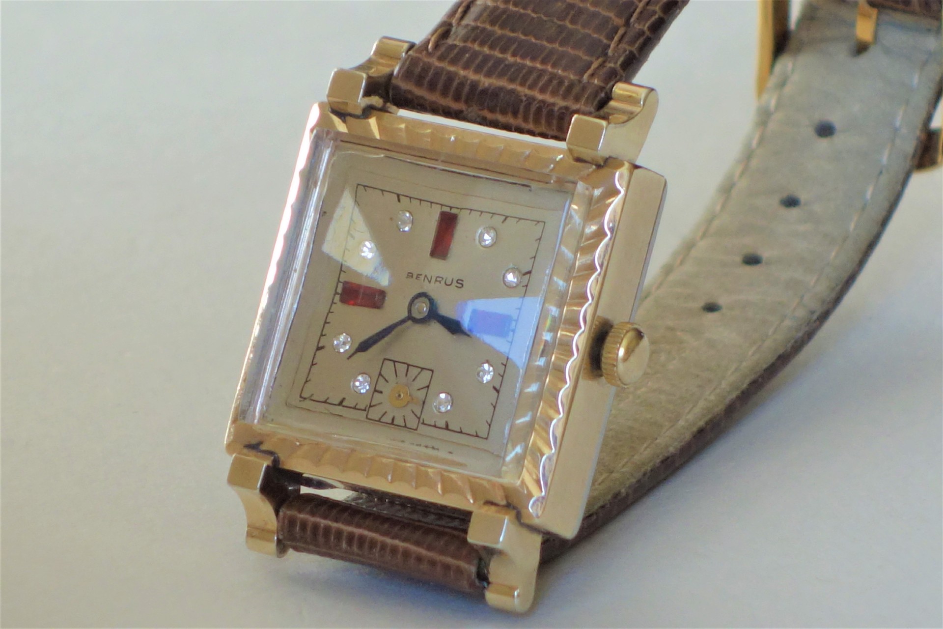 Eckige Armbanduhr mit goldfarbenem Gehäuse, beigem Zifferblatt, braunem Lederarmband und Schmucksteinen als Indizes.