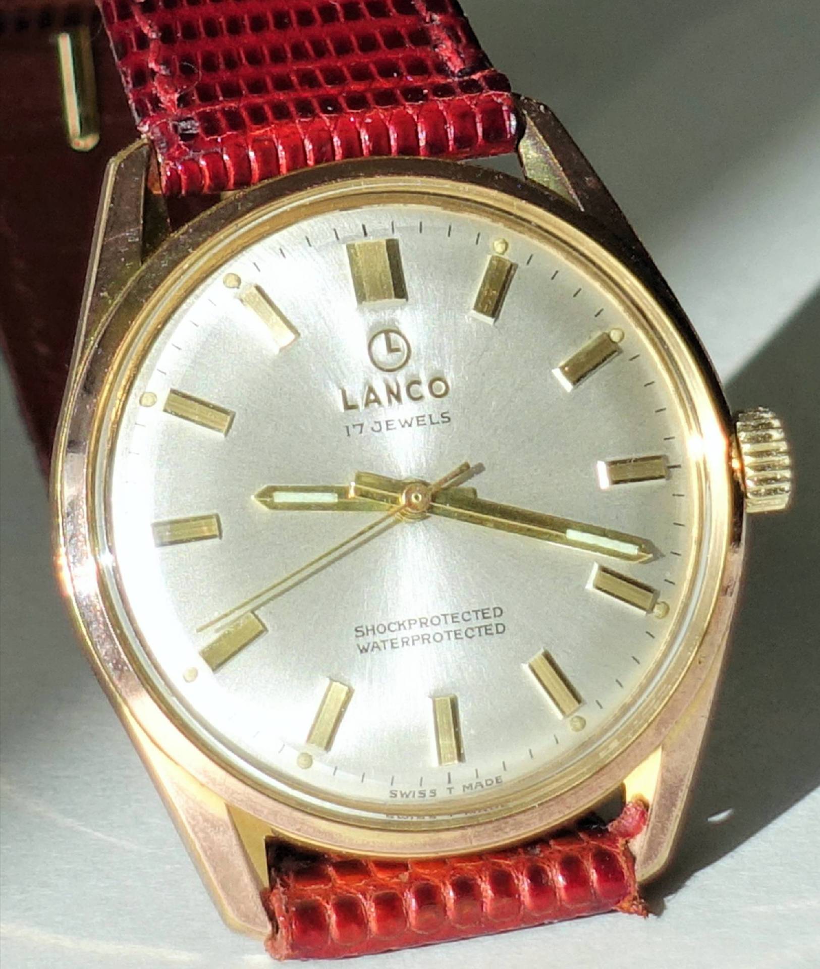 Goldfarbene Armbanduhr mit silberfarbenem Zifferblatt und rotem Armband.