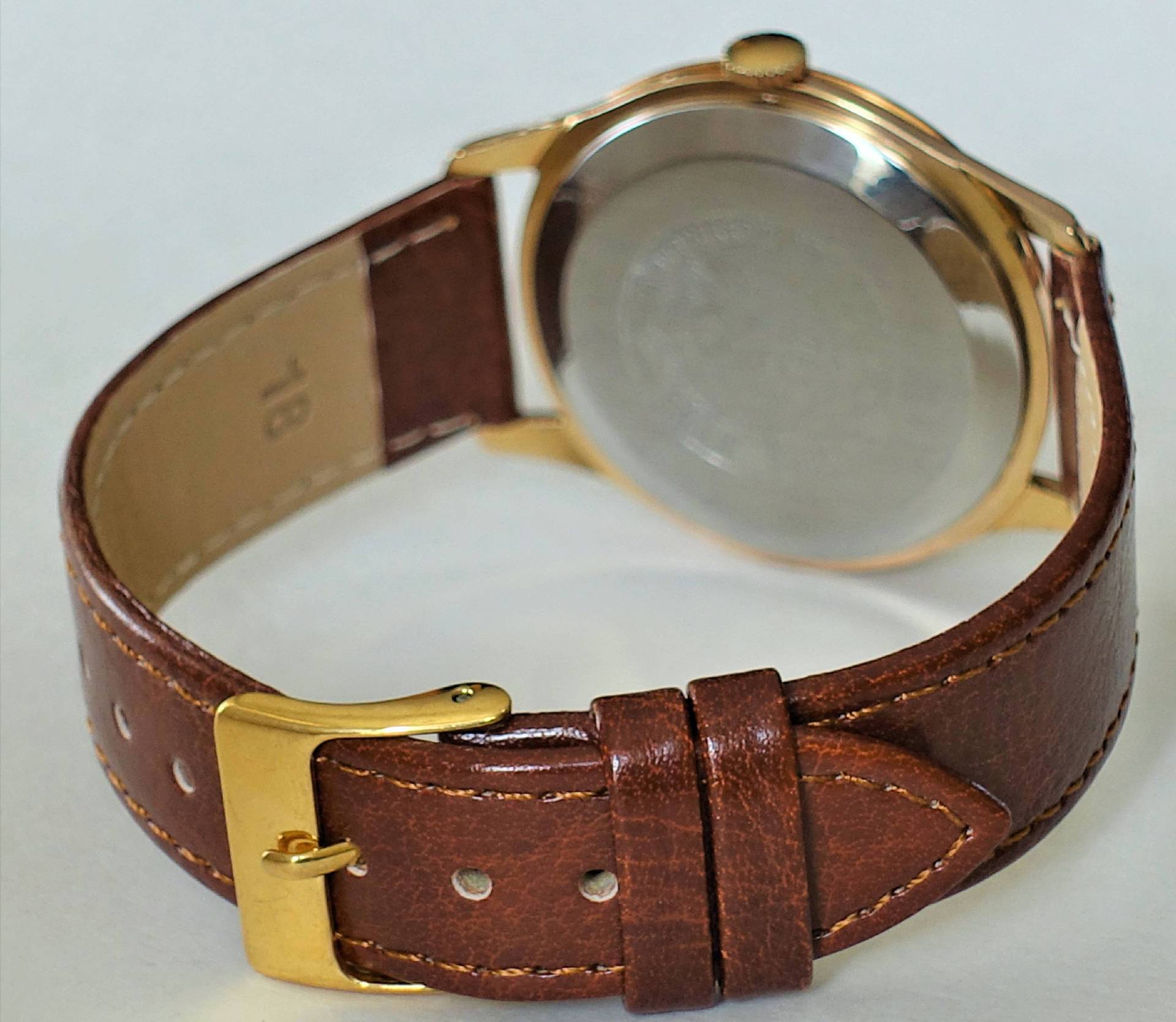 Eine Armbanduhr mit braunem Lederarmband, goldfarbenem Gehäuse und silbernem Gehäuseboden ist von hinten zu sehen.