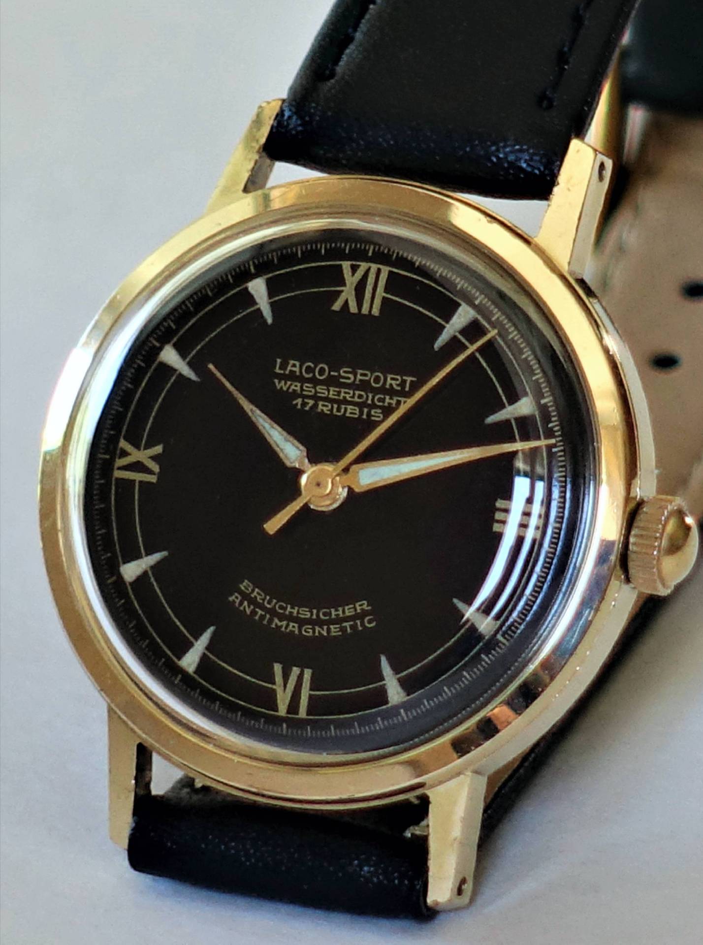 Schwarze Armbanduhr mit goldfarbenem Gehäuse und schwarzem Lederarmband, Zifferblatt mit römischen Zahlen.