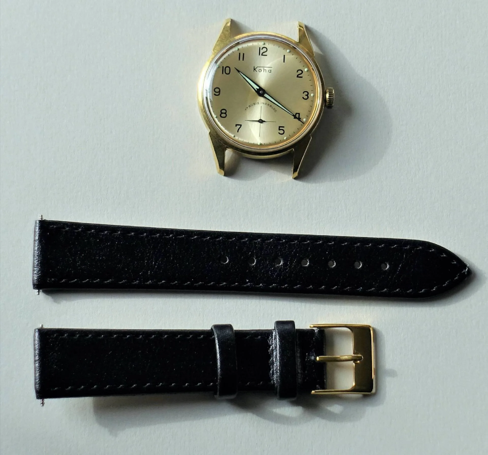 Goldfarbene Armbanduhr mit hellem Zifferblatt und schwarzem Lederarmband mit goldfarbener Schließe.