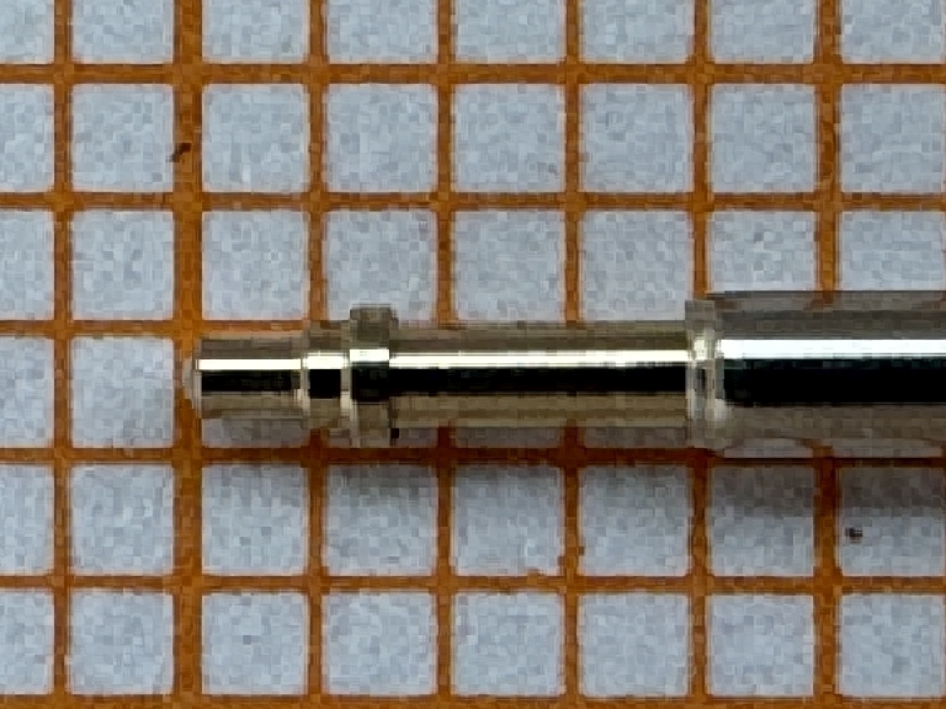 Metallene Aufzugswelle für Armbanduhren, liegend auf kariertem Hintergrund.