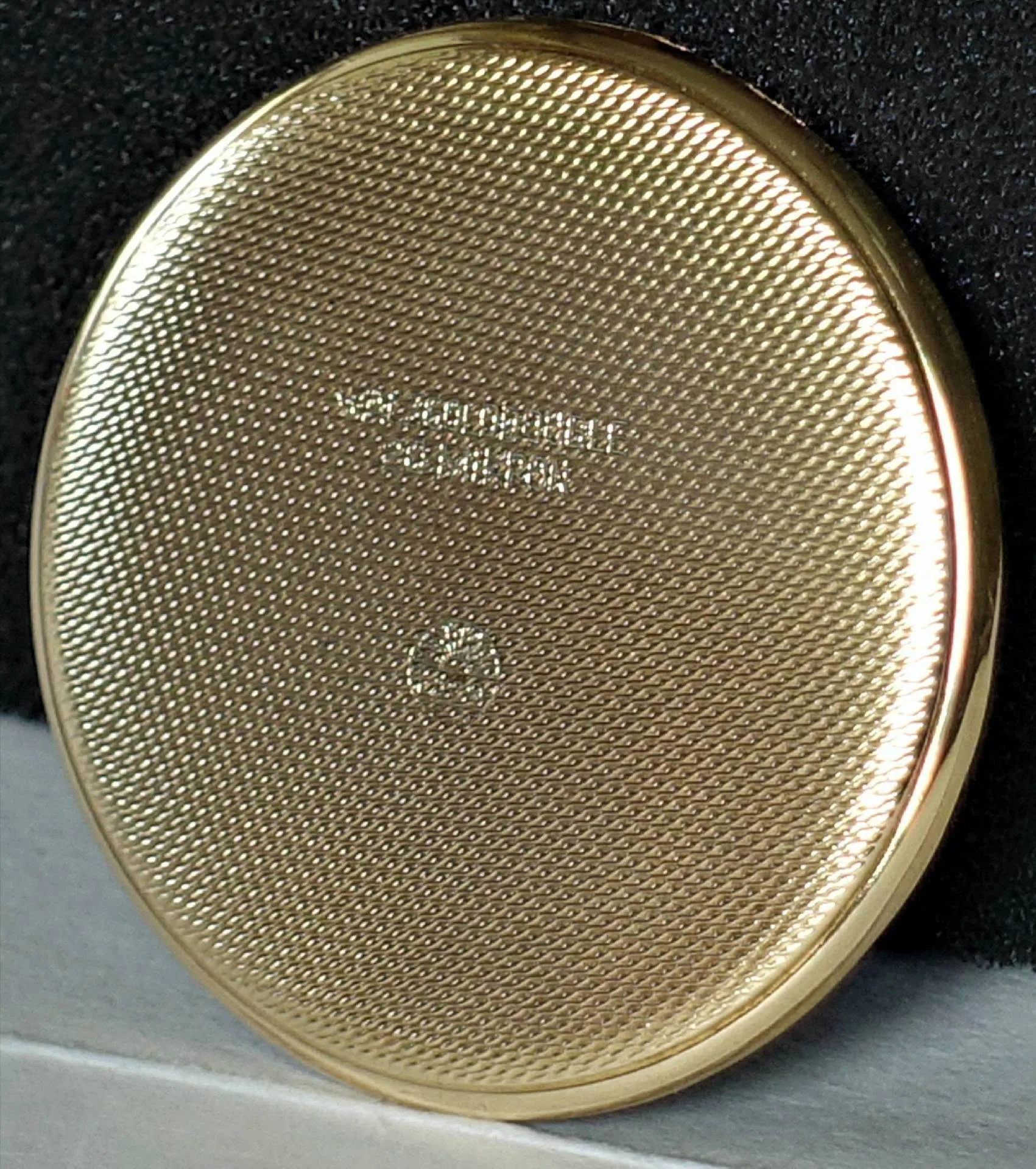 Goldfarbener Uhrenboden mit Muster Rückseite eines goldfarbenen, fein gemusterten Uhrenbodens vor dunklem Hintergrund.