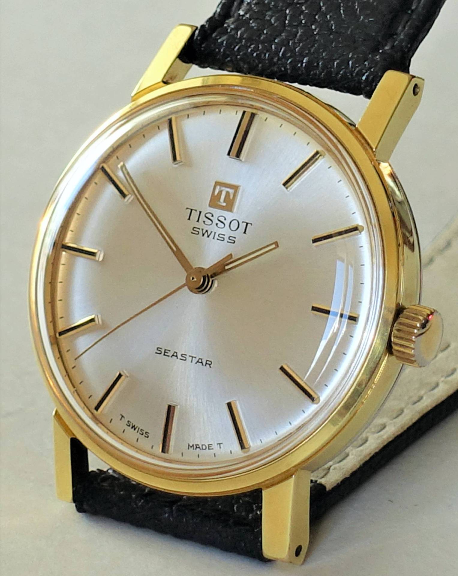 Klassische Armbanduhr mit goldfarbenem Gehäuse, silberfarbenem Zifferblatt und schwarzem Armband.