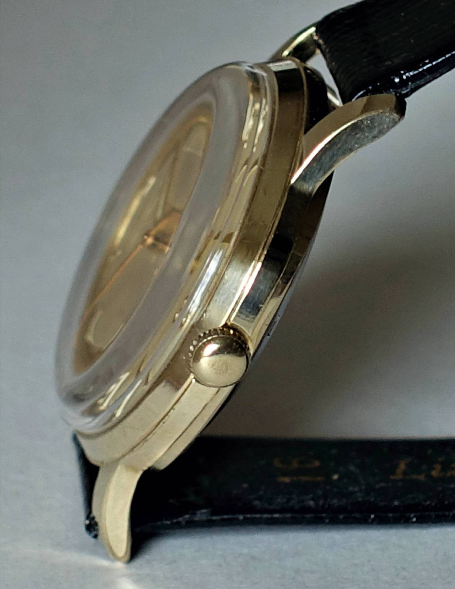 Goldfarbene Armbanduhr mit schwarzem Armband und seitlicher Krone.