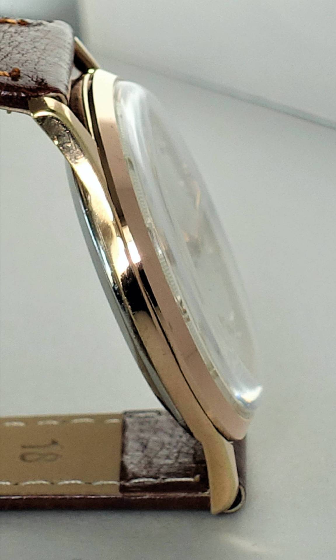 Seitenansicht eines flachen Uhrgehäuses in goldener Farbe mit braunem Lederarmband und gewölbtem Glas.