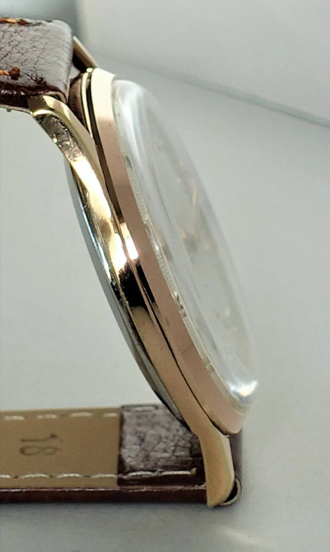 Seitenansicht eines flachen Uhrgehäuses in goldener Farbe mit braunem Lederarmband und gewölbtem Glas.