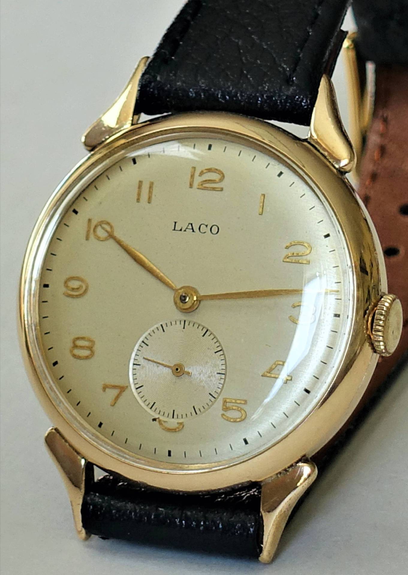 Goldfarbene Armbanduhr mit hellem Zifferblatt, schwarzen Zahlen und schwarzem Lederarmband.