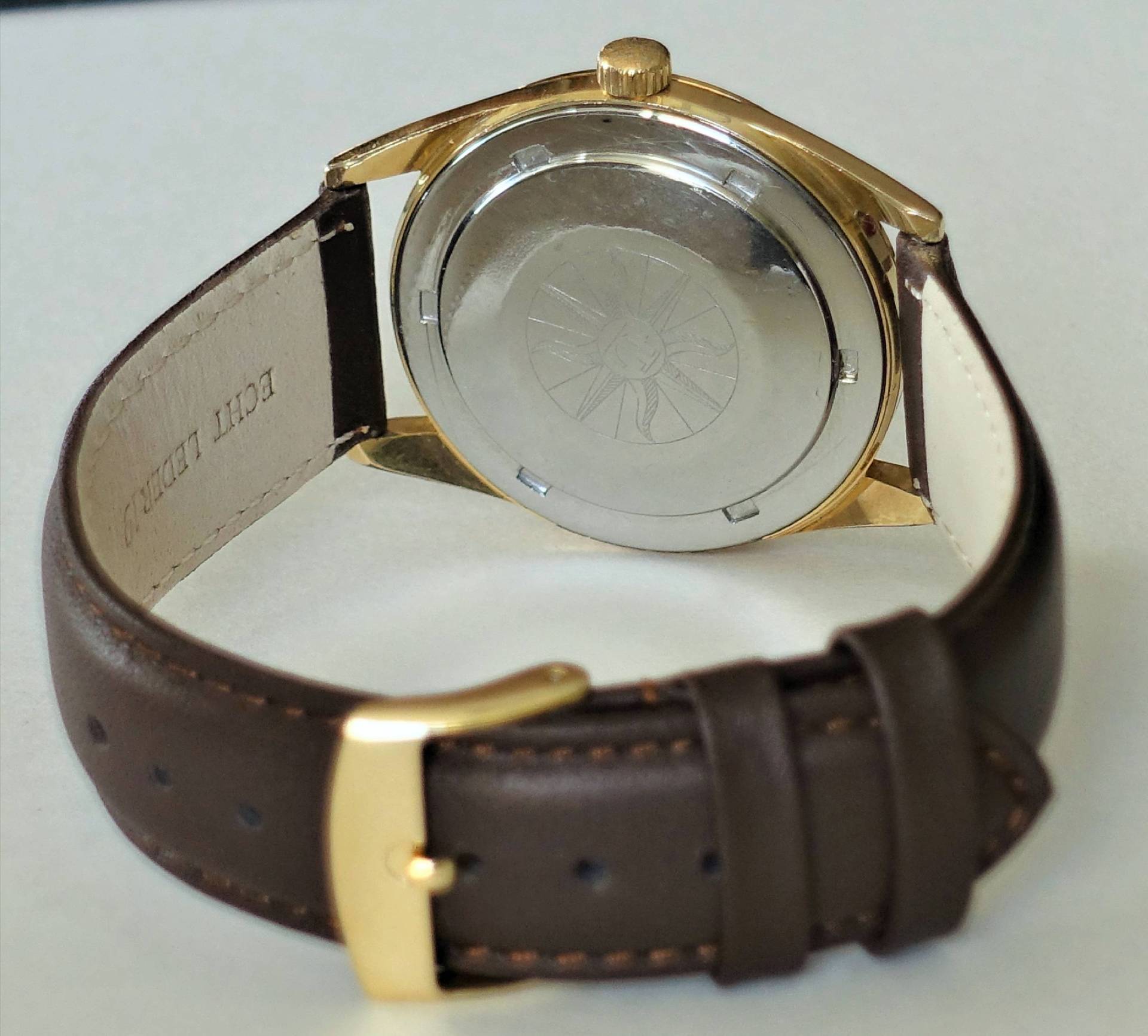 Armbanduhr mit goldfarbenem Gehäuse, braunem Lederarmband und silberfarbener Rückseite.