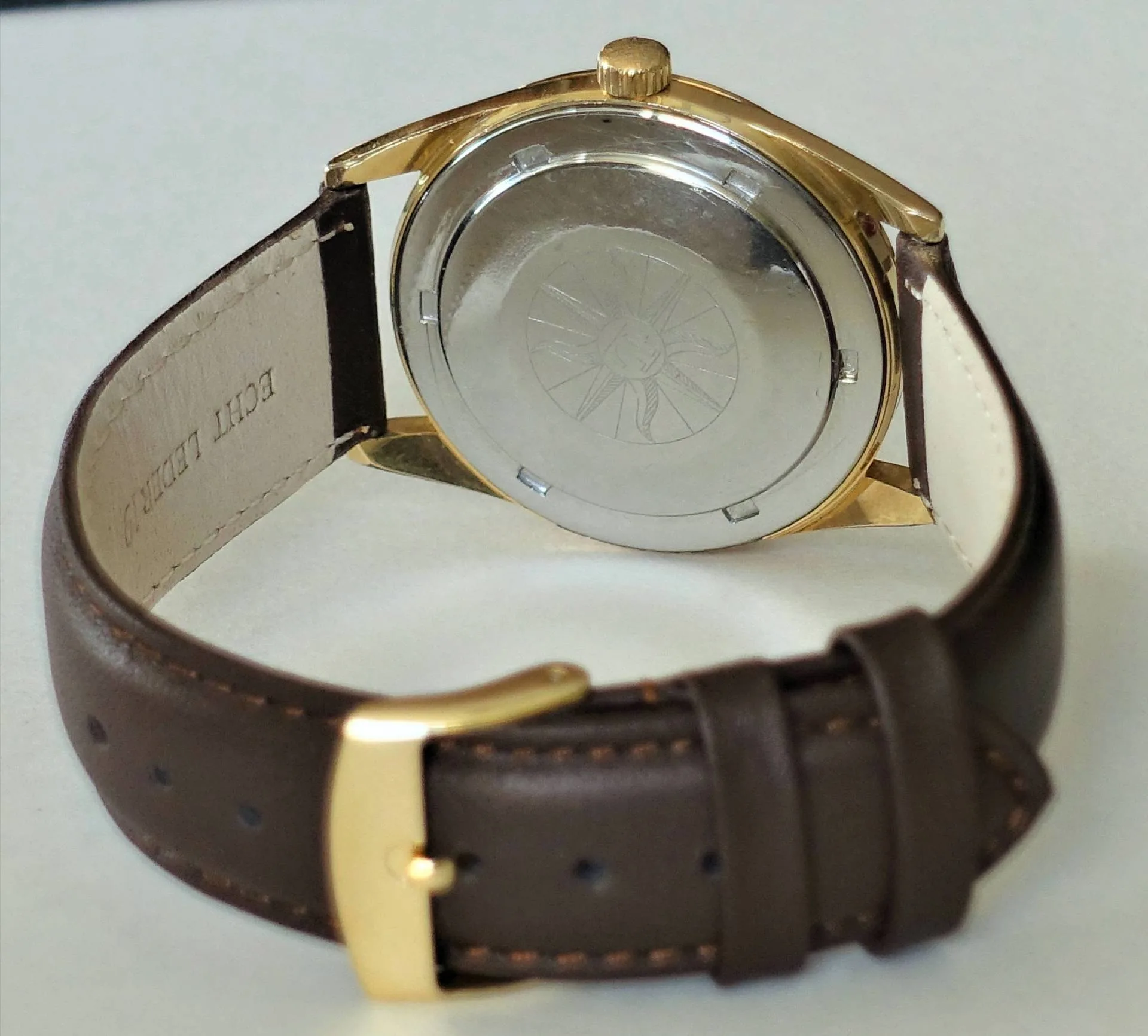 Armbanduhr mit goldfarbenem Gehäuse, braunem Lederarmband und silberfarbener Rückseite.