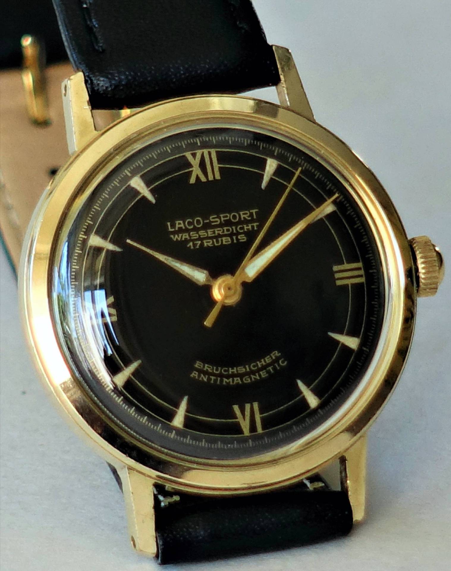 Schwarze Armbanduhr mit goldfarbenem Gehäuse und schwarzem Lederarmband, Zifferblatt mit römischen Zahlen und goldenen Zeigern.