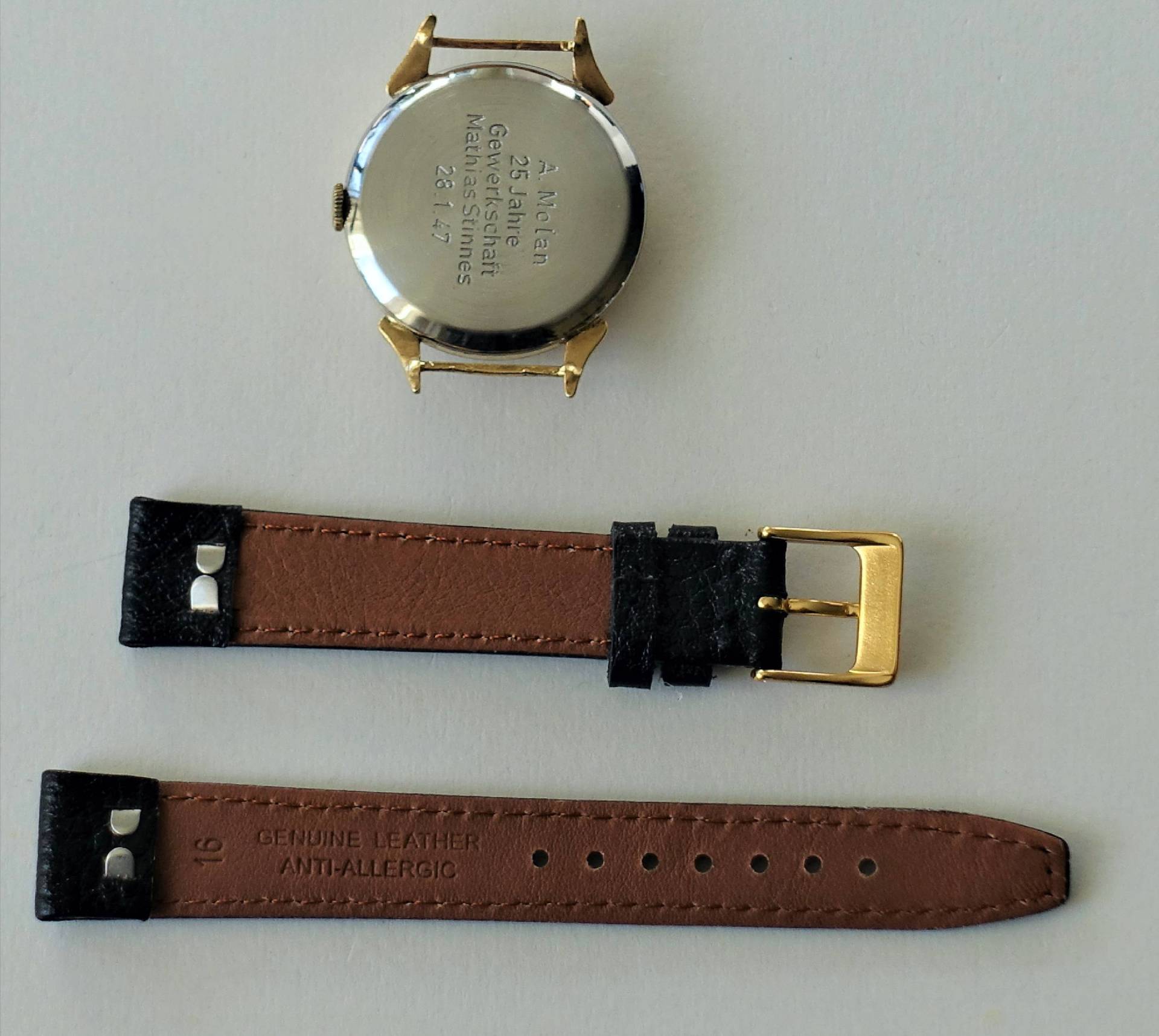 Gehäuse einer Armbanduhr in goldfarbener Optik mit graviertem Metallboden, daneben ein zweiteiliges braun-schwarzes Lederarmband mit goldfarbener Schließe.