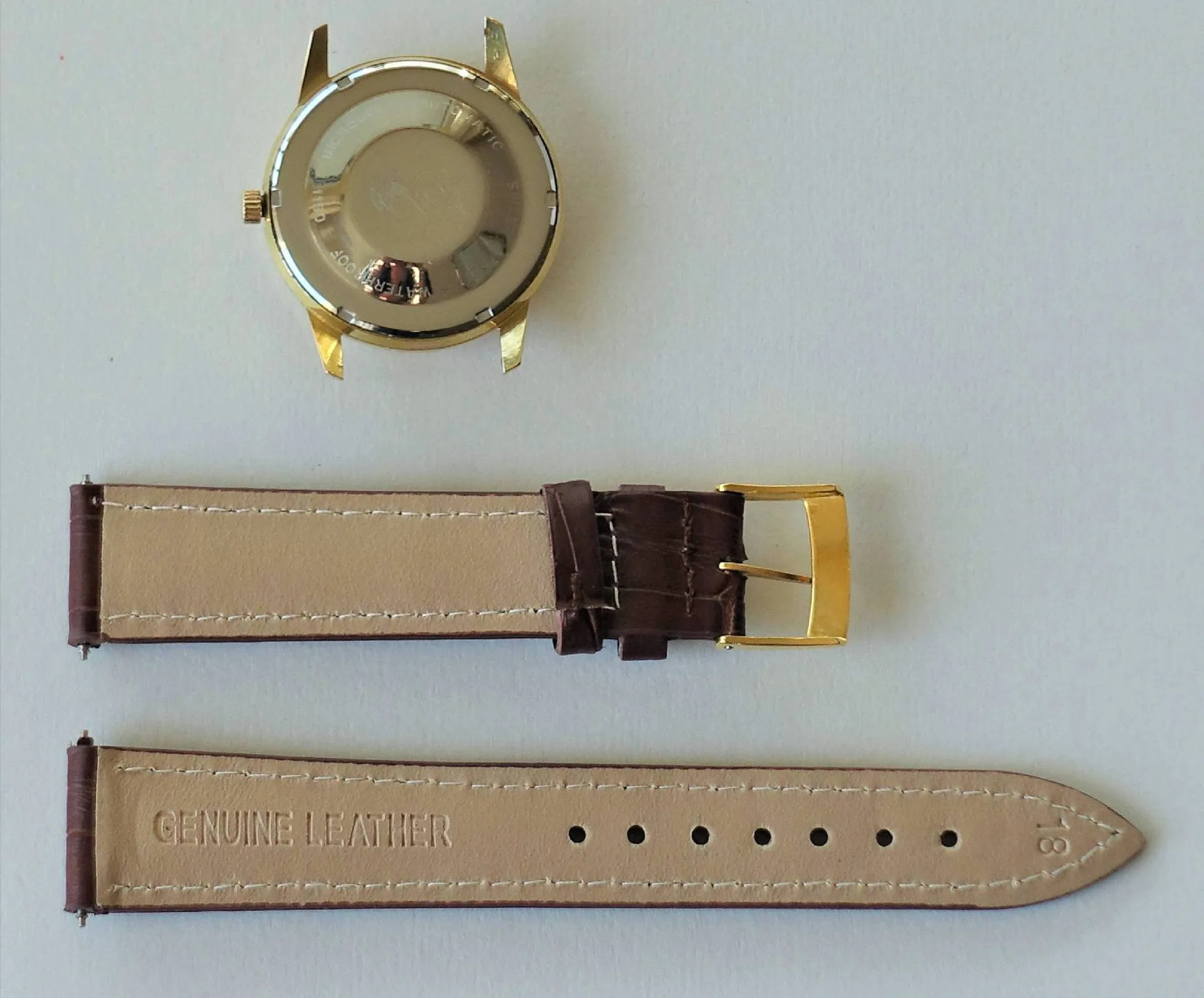 Ein goldfarbenes Uhrengehäuse und ein zweiteiliges braunes Lederarmband mit goldfarbener Schließe liegen nebeneinander auf einer hellen Oberfläche.