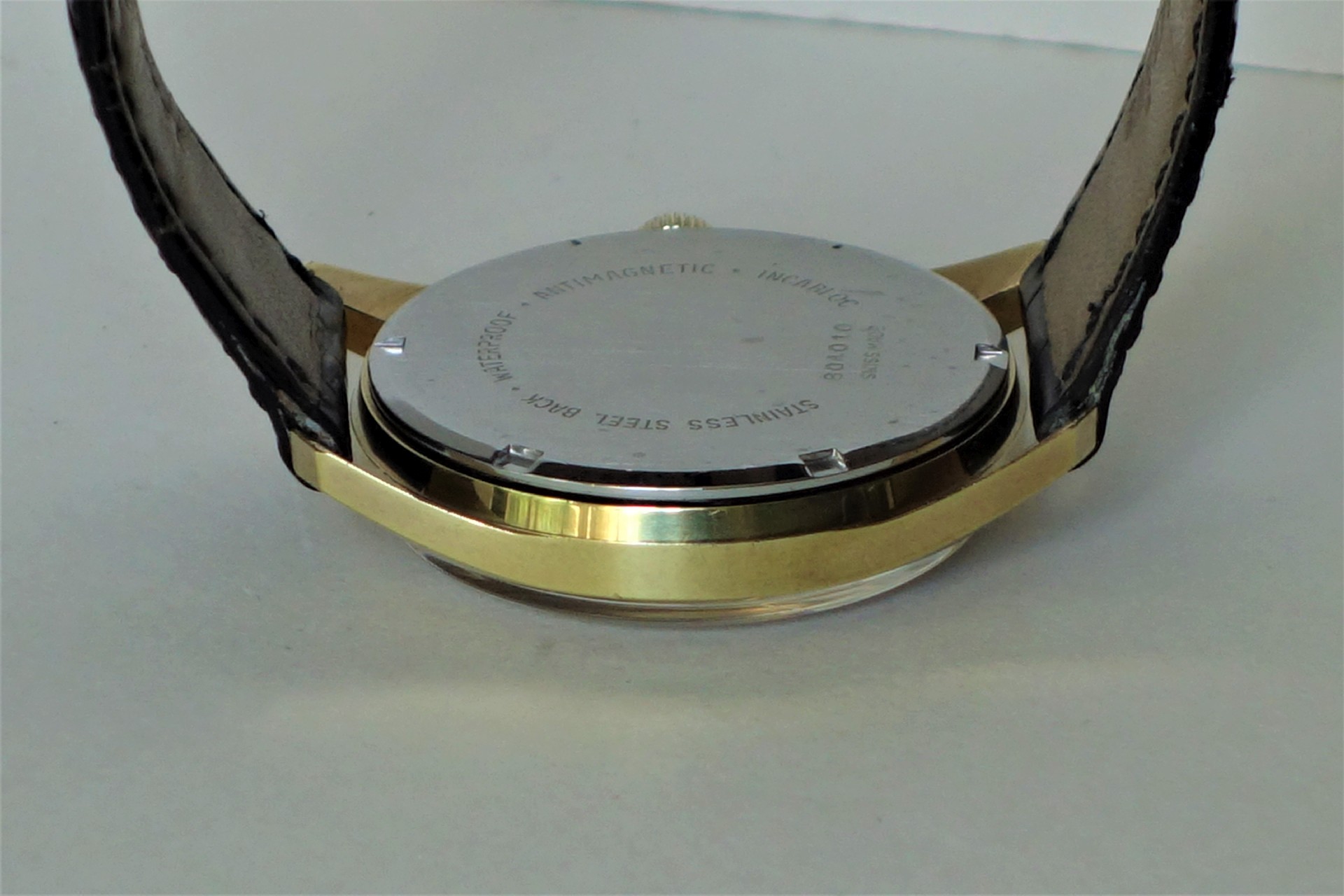 Runder Uhrenboden aus Edelstahl an einer Armbanduhr mit goldfarbenem Gehäuse und schwarzem Lederarmband.