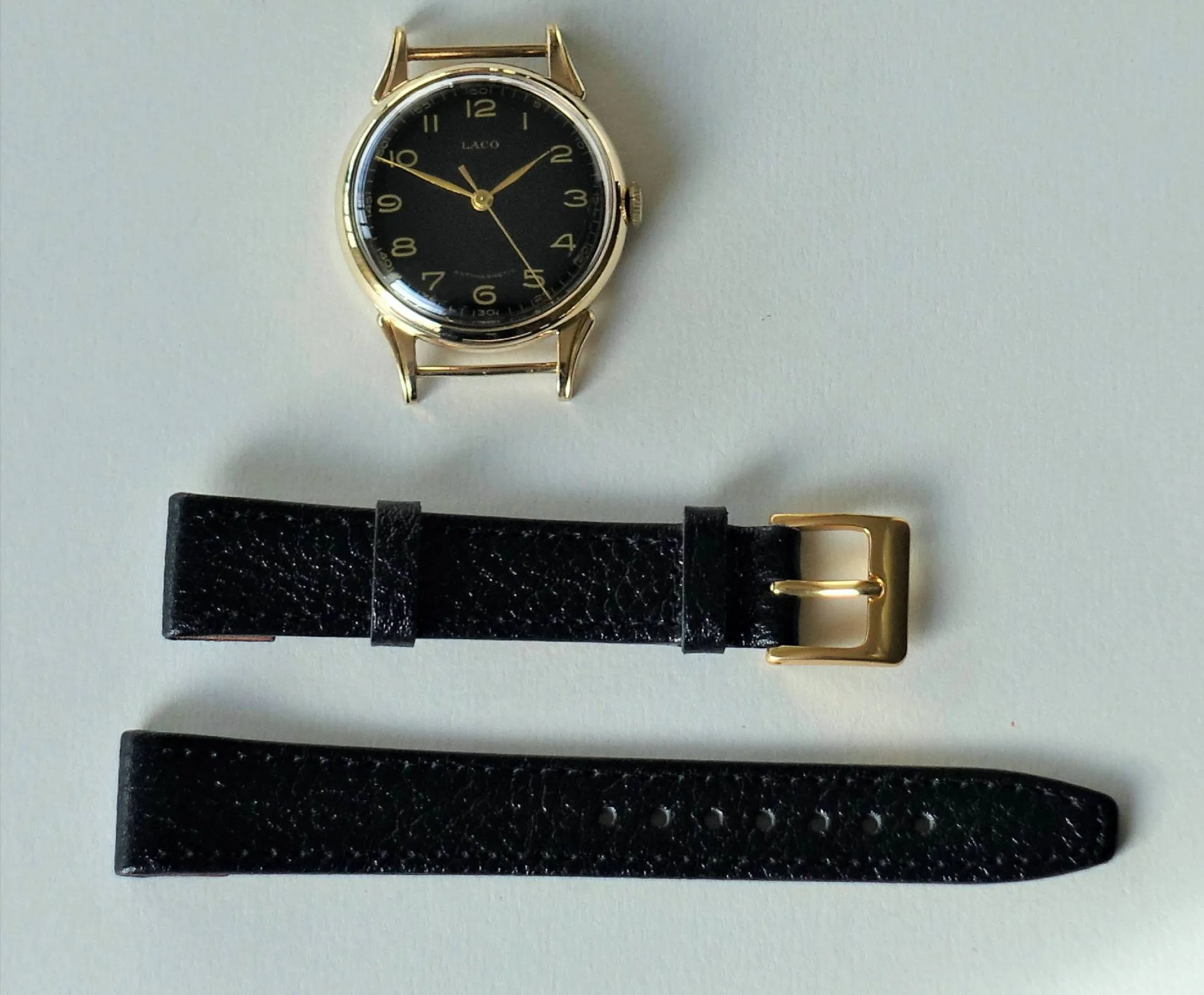 Eine Armbanduhr mit schwarzem Zifferblatt und schwarzem Lederarmband liegt in Einzelteilen auf hellem Untergrund.