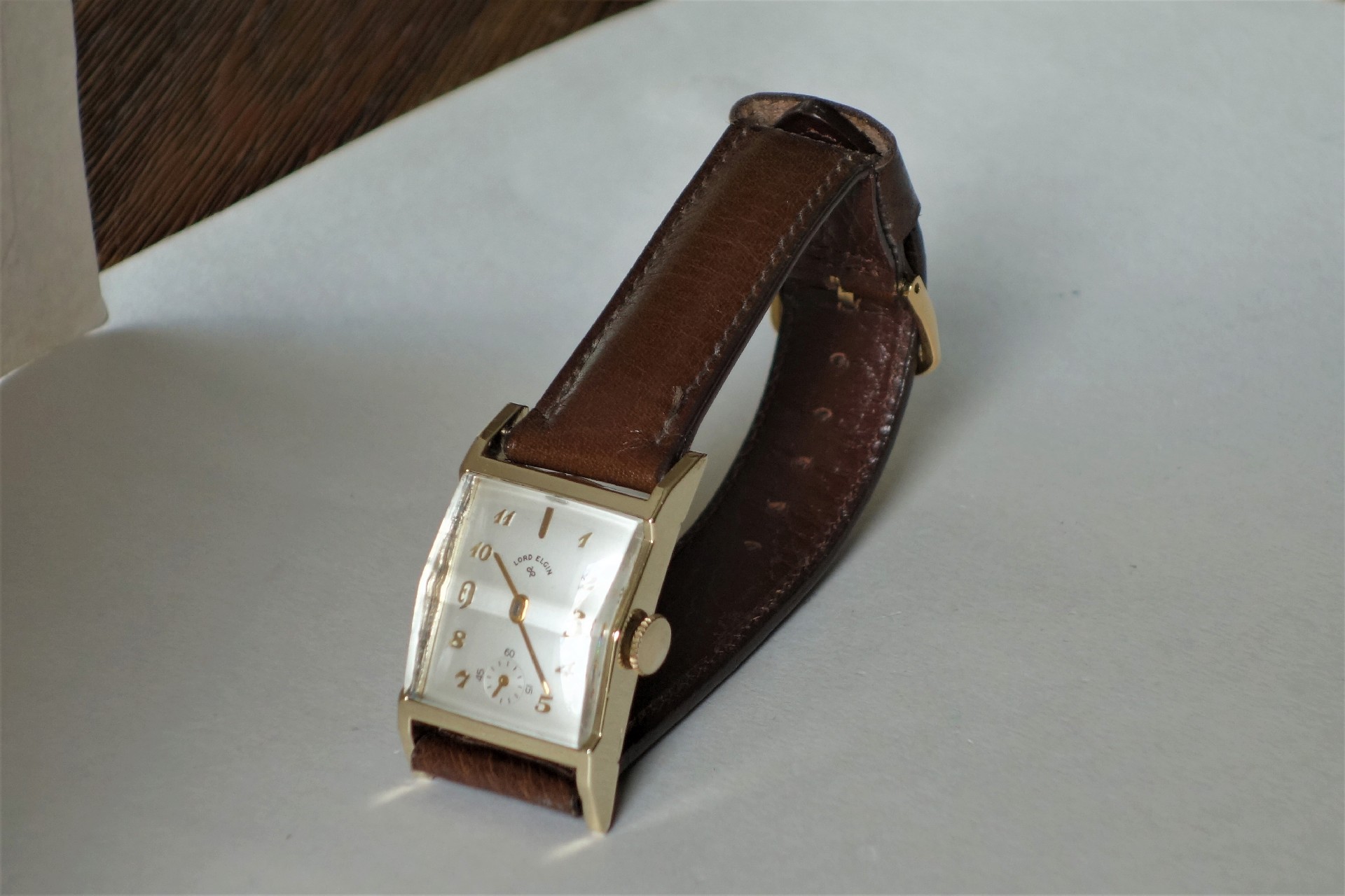Rechteckige Armbanduhr mit braunem Armband und hellem Zifferblatt.
