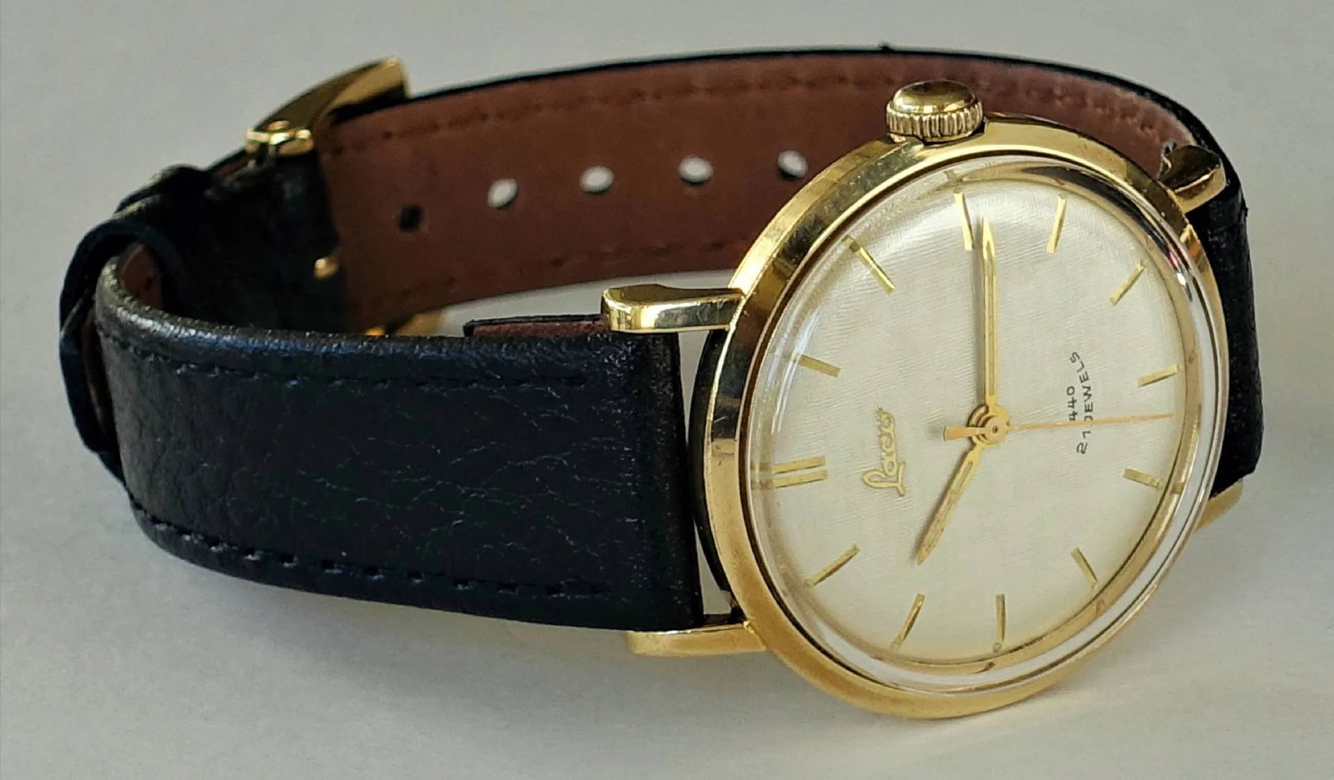 Analoge Armbanduhr mit goldfarbenem Gehäuse, hellem Zifferblatt und schwarzem Lederarmband.