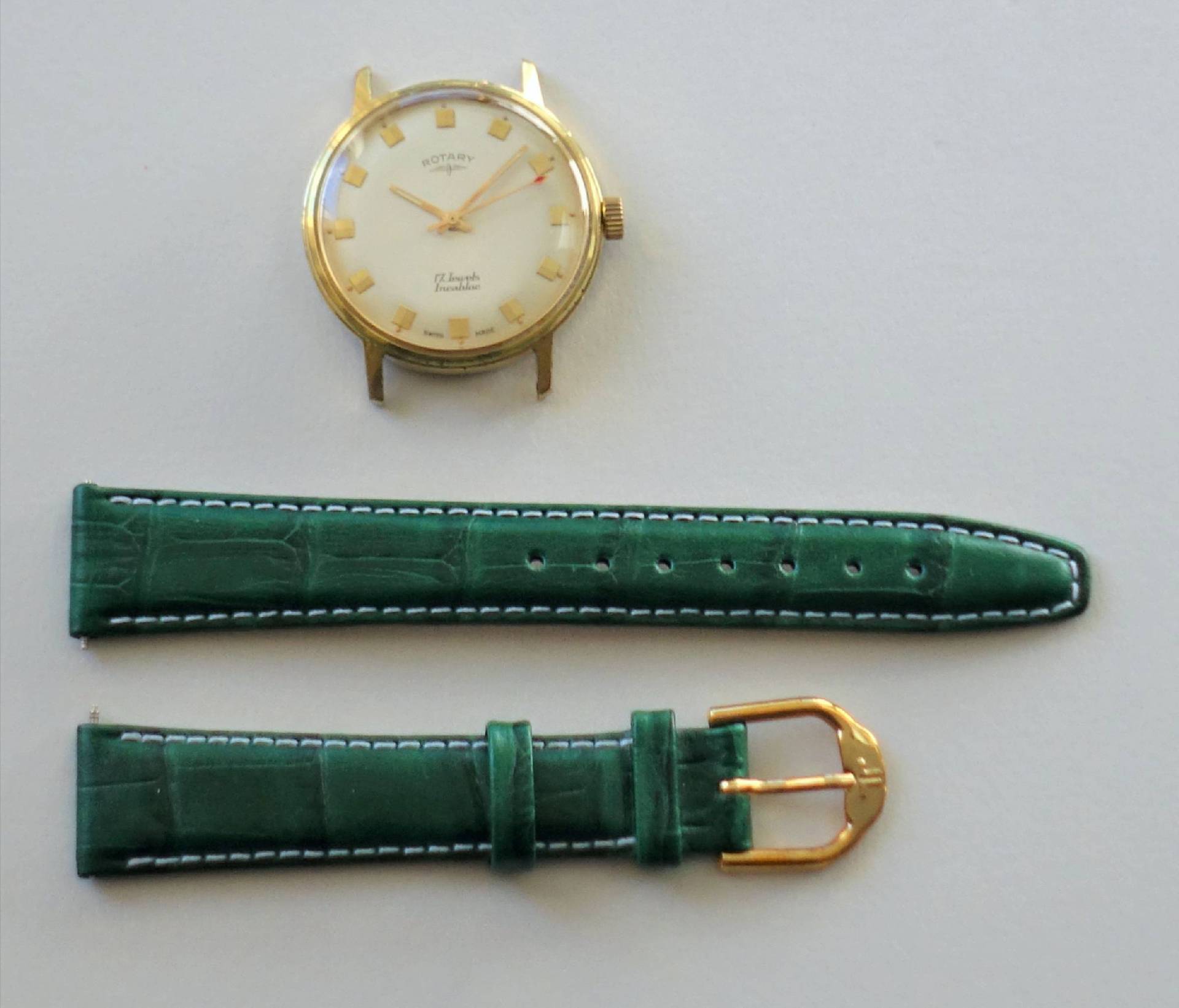 Goldfarbene Armbanduhr mit weißem Zifferblatt und grünem Armband mit goldfarbener Schließe.