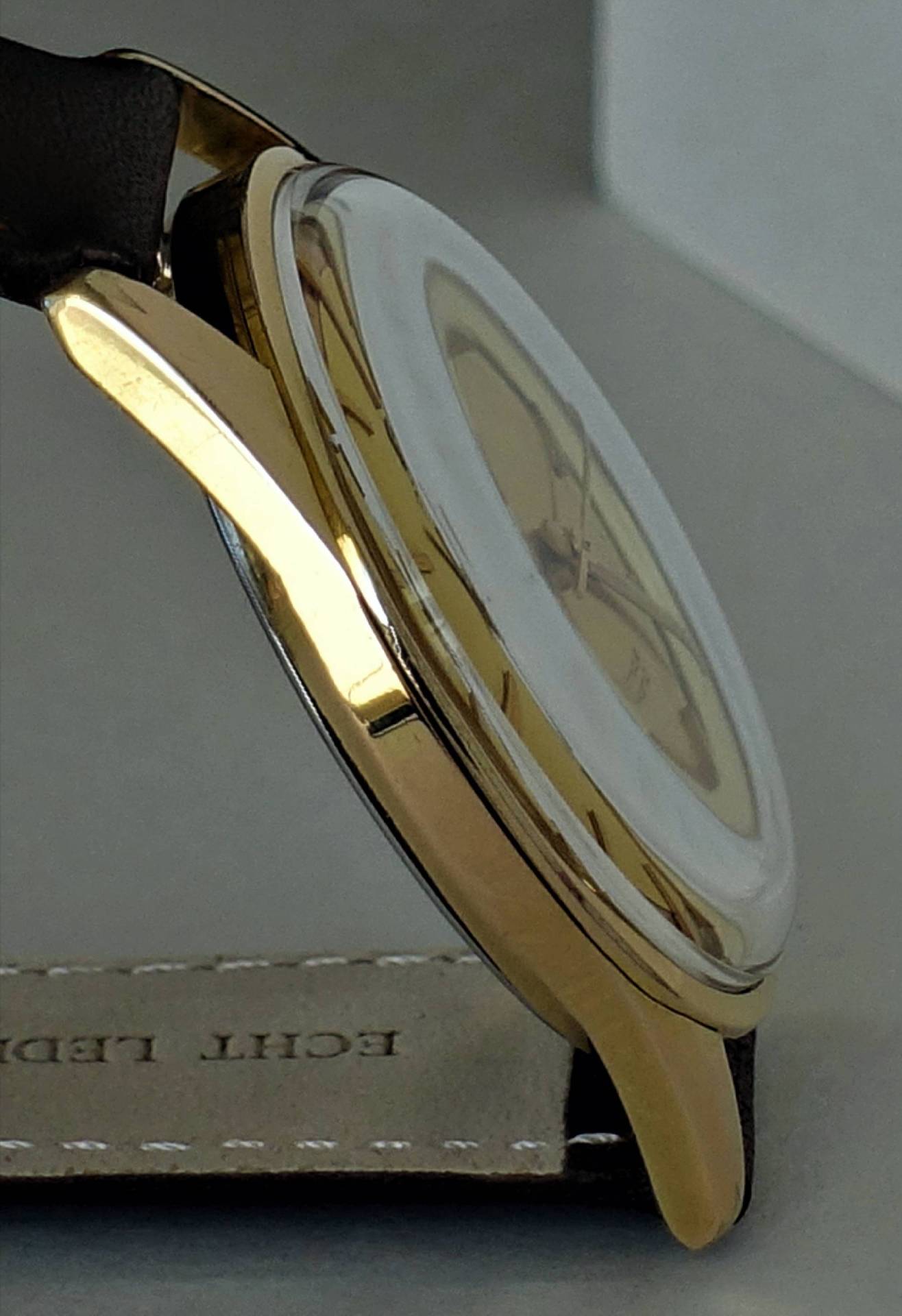 Goldfarbene Armbanduhr mit hellem Zifferblatt und dunklem Armband in Seitenansicht.