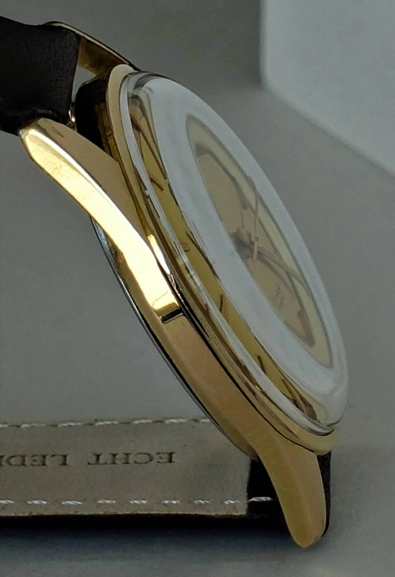 Goldfarbene Armbanduhr mit hellem Zifferblatt und dunklem Armband in Seitenansicht.