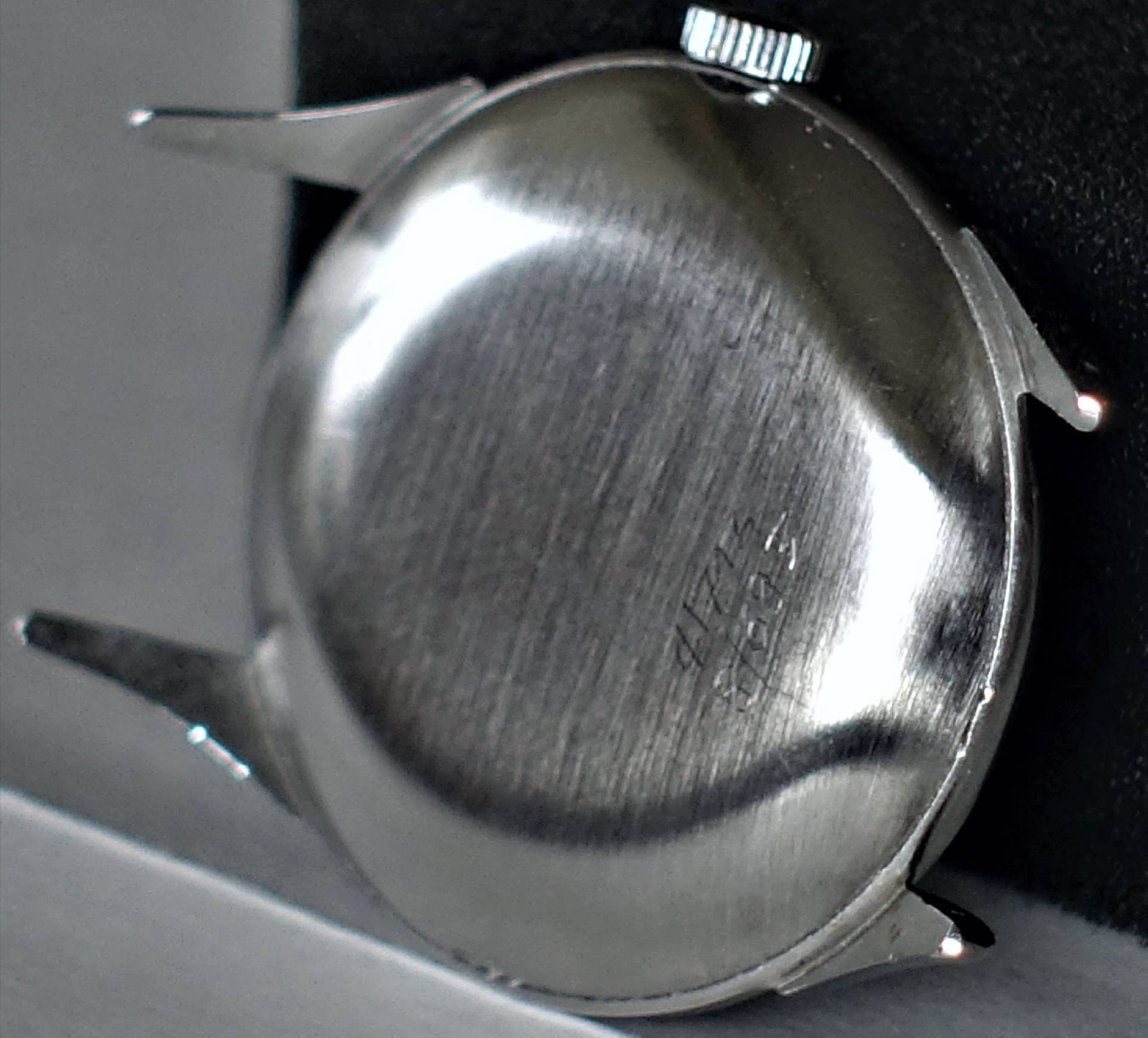 Rückseite eines silberfarbenen Uhrgehäuses mit sichtbarer Krone und Gravur.