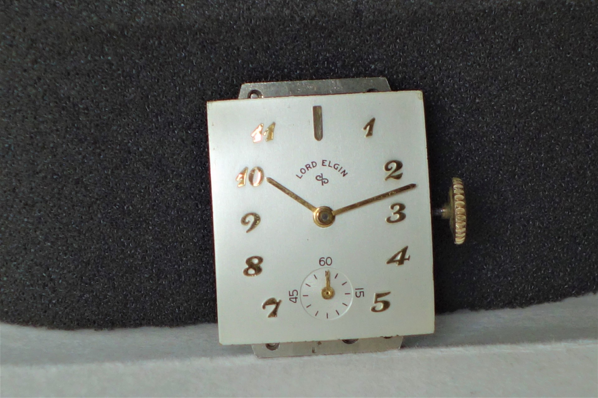 Rechteckiges Uhrwerk mit silberfarbenem Zifferblatt und goldfarbenen Zeigern, kleine Sekunde bei sechs Uhr.
