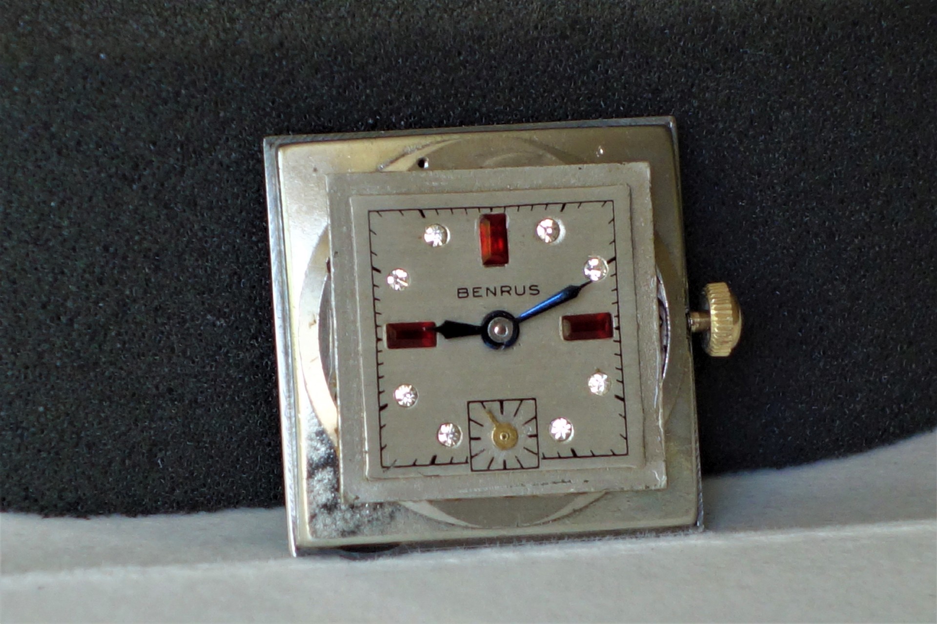 Mechanisches Uhrwerk mit quadratischem silberfarbenem Zifferblatt, roten und weißen Indizes sowie blauen Zeigern.