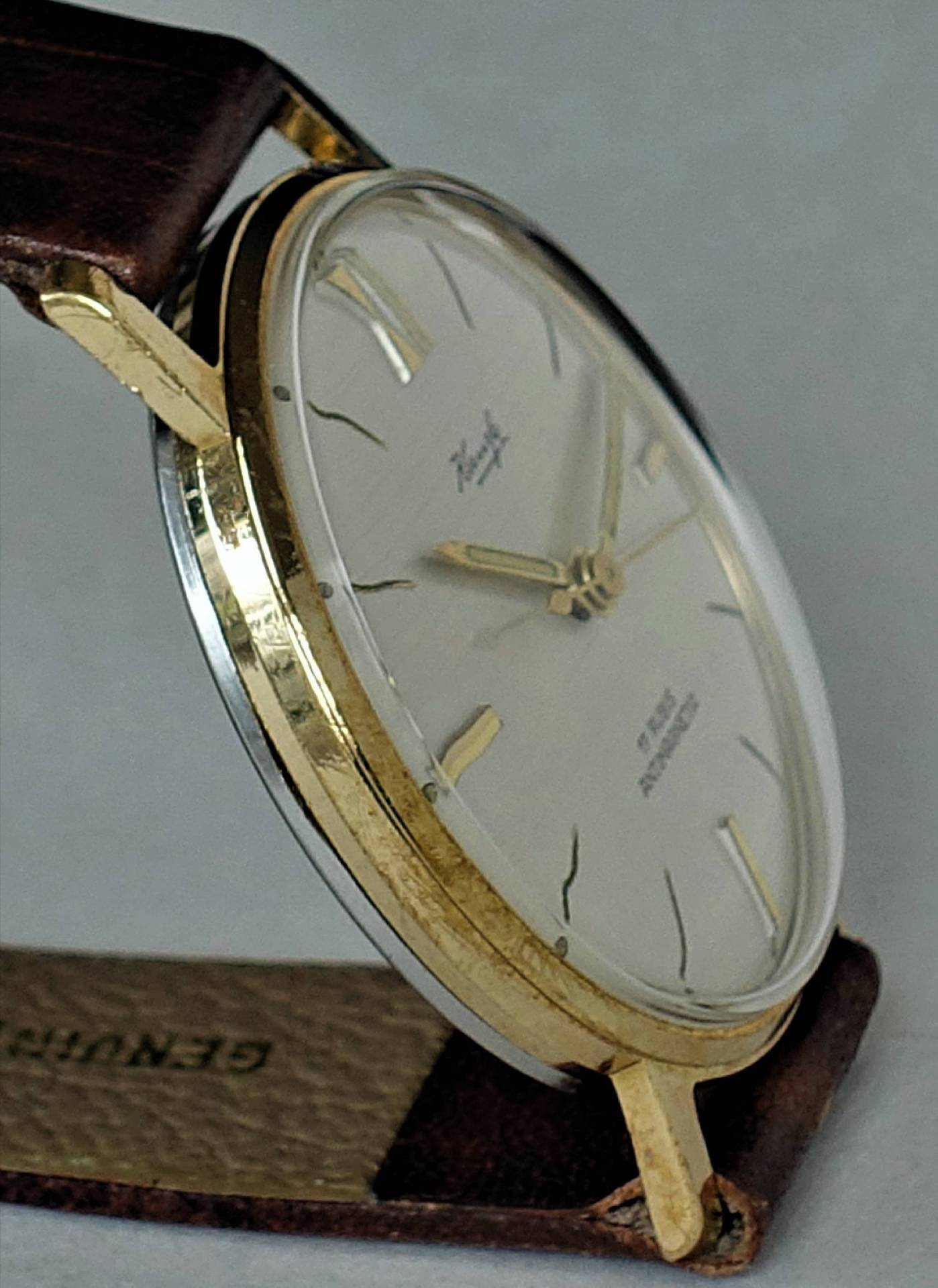 Flache Armbanduhr mit goldfarbenem Gehäuse, hellem Zifferblatt und braunem Armband.